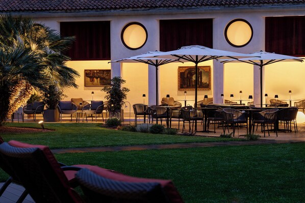Un área de comedor al aire libre con mesas y sillas, rodeada de palmeras y un edificio con grandes ventanales.