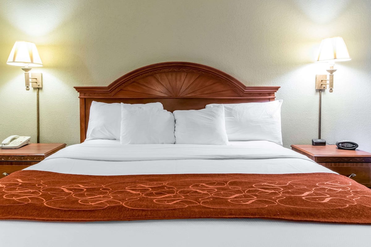 Un lit bien fait, avec du linge de lit blanc et une couverture marron, flanqué de deux tables de chevet sur lesquelles reposent des lampes et un téléphone.
