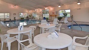 A poolside area with multiple white plastic tables and chairs, a pool, and a view of the outdoors through large windows.