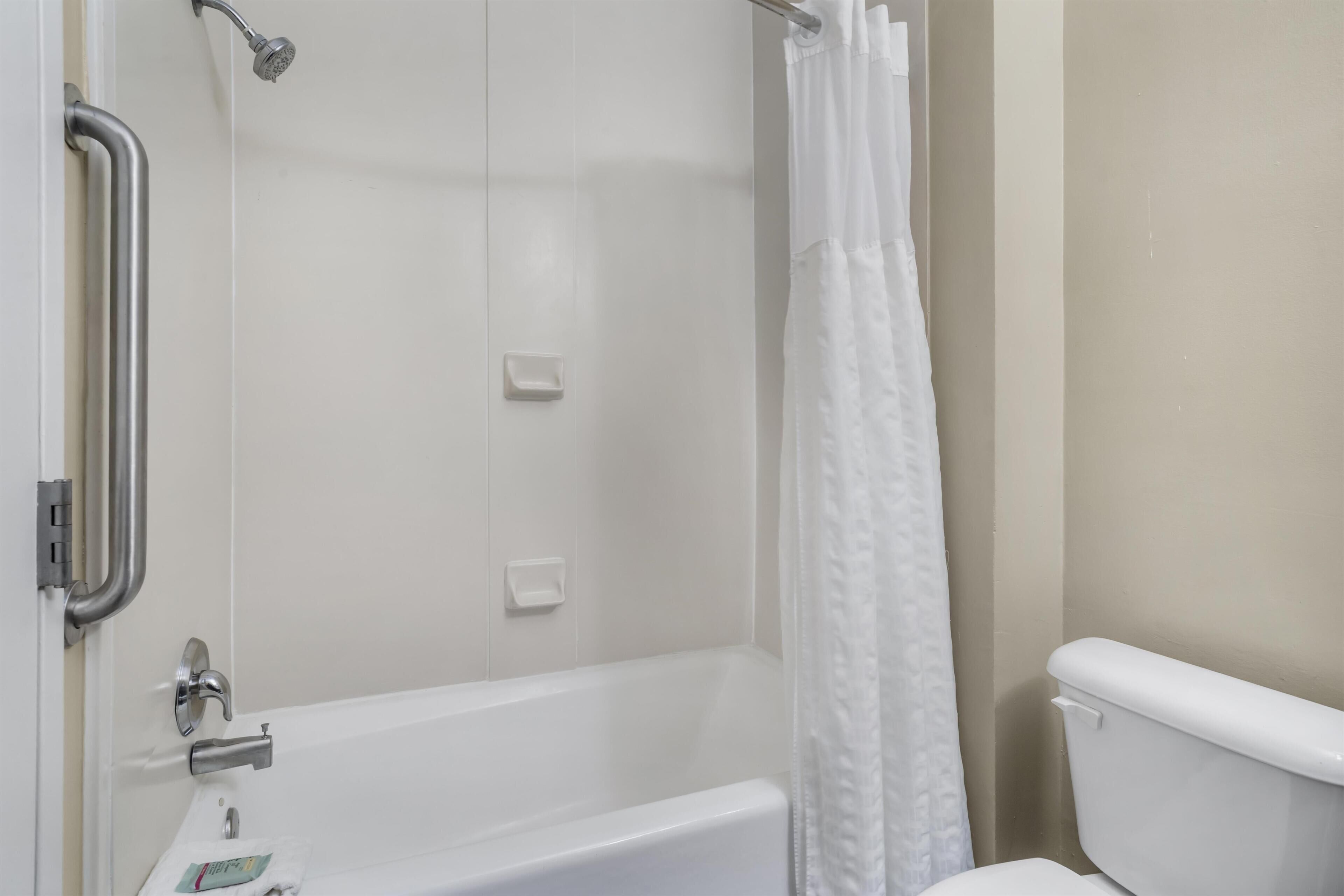 Une salle de bain avec une baignoire blanche, des toilettes, une douche avec un rideau blanc et une barre d’appui.