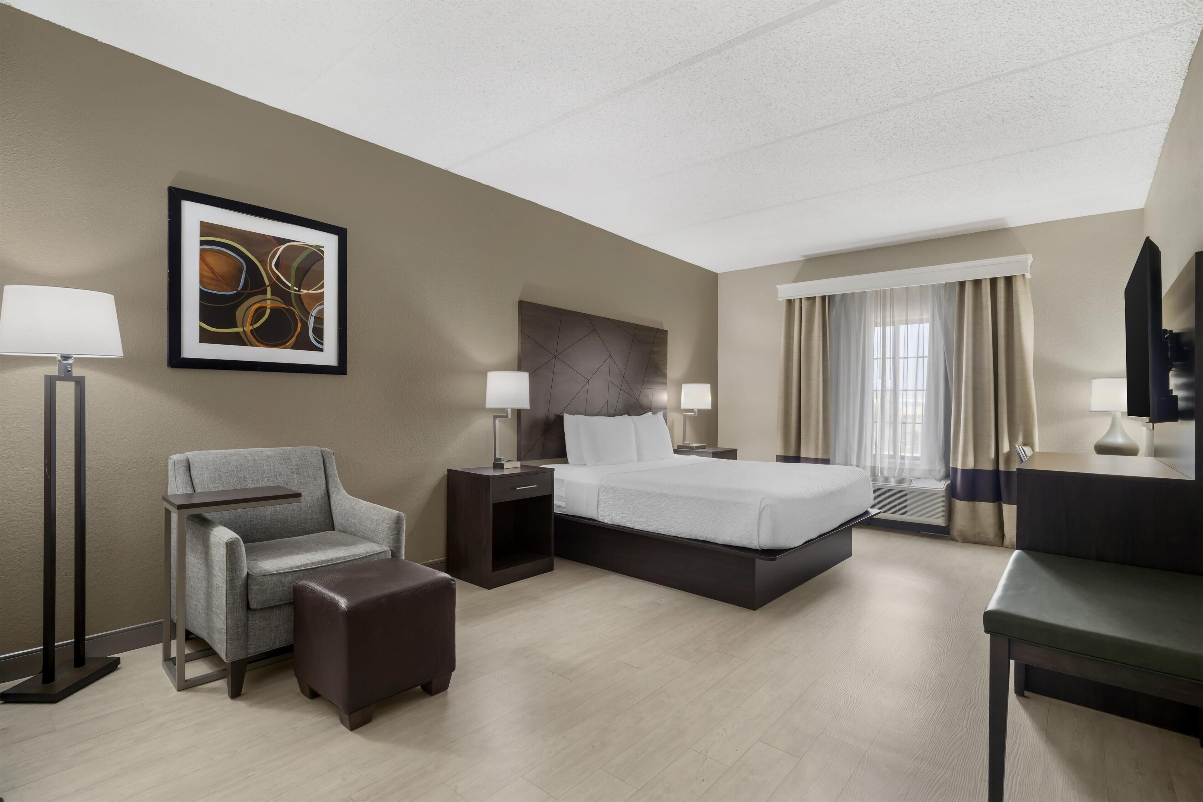 A hotel room with a bed, a chair, a desk, a lamp, a nightstand, and a picture on the wall.