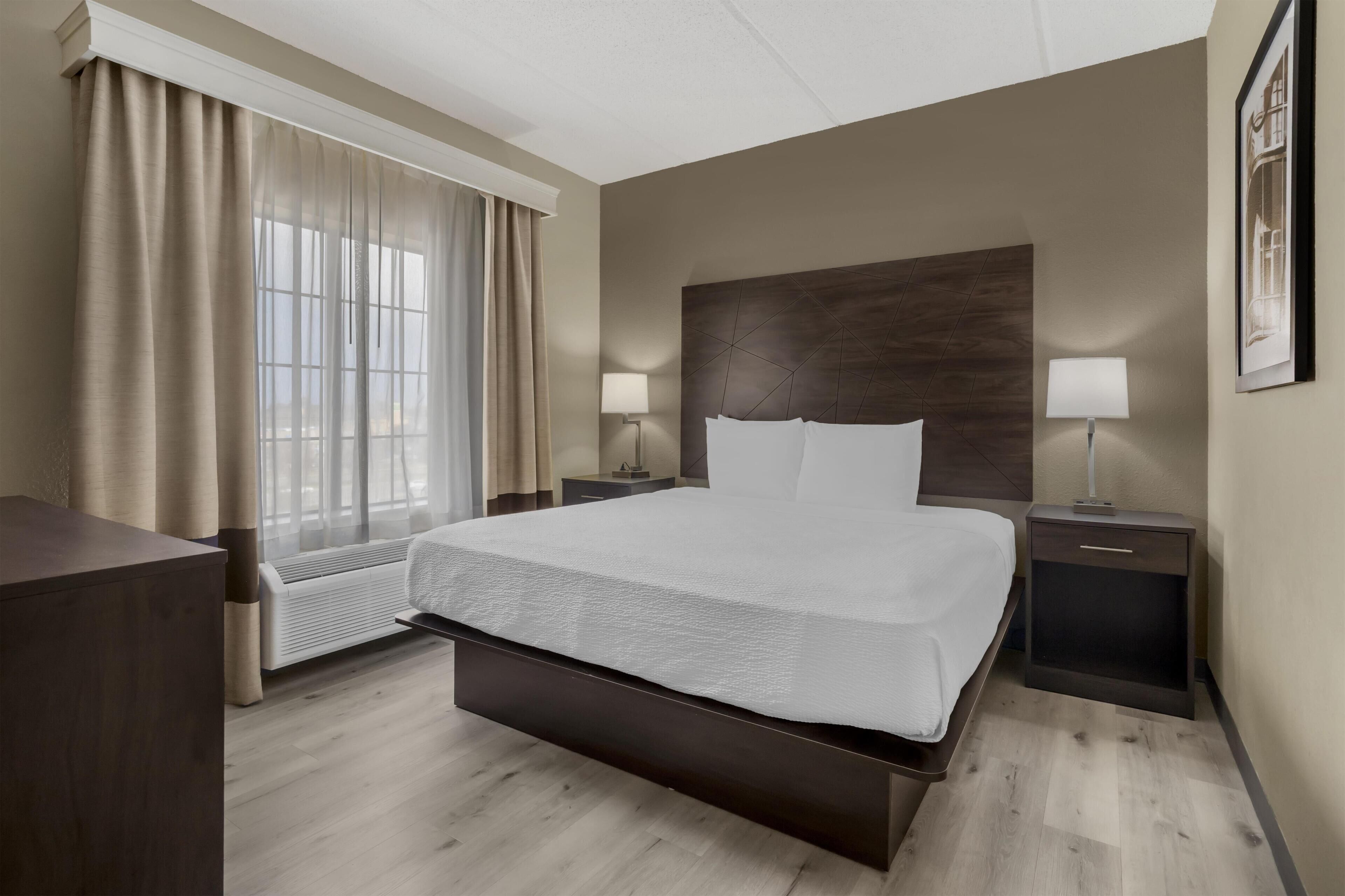 A hotel room with a large bed, bedside lamps, a nightstand, a window with curtains, and a framed picture on the wall.