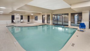 An indoor swimming pool with clear water, a lounge area with chairs, and a view of the outside through large windows.