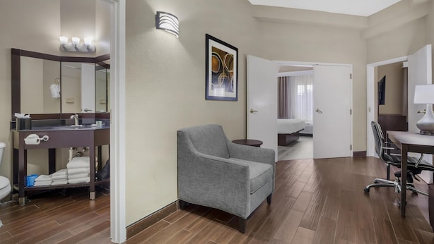 A hotel room with a bathroom, a gray armchair, a desk, and a painting on the wall.