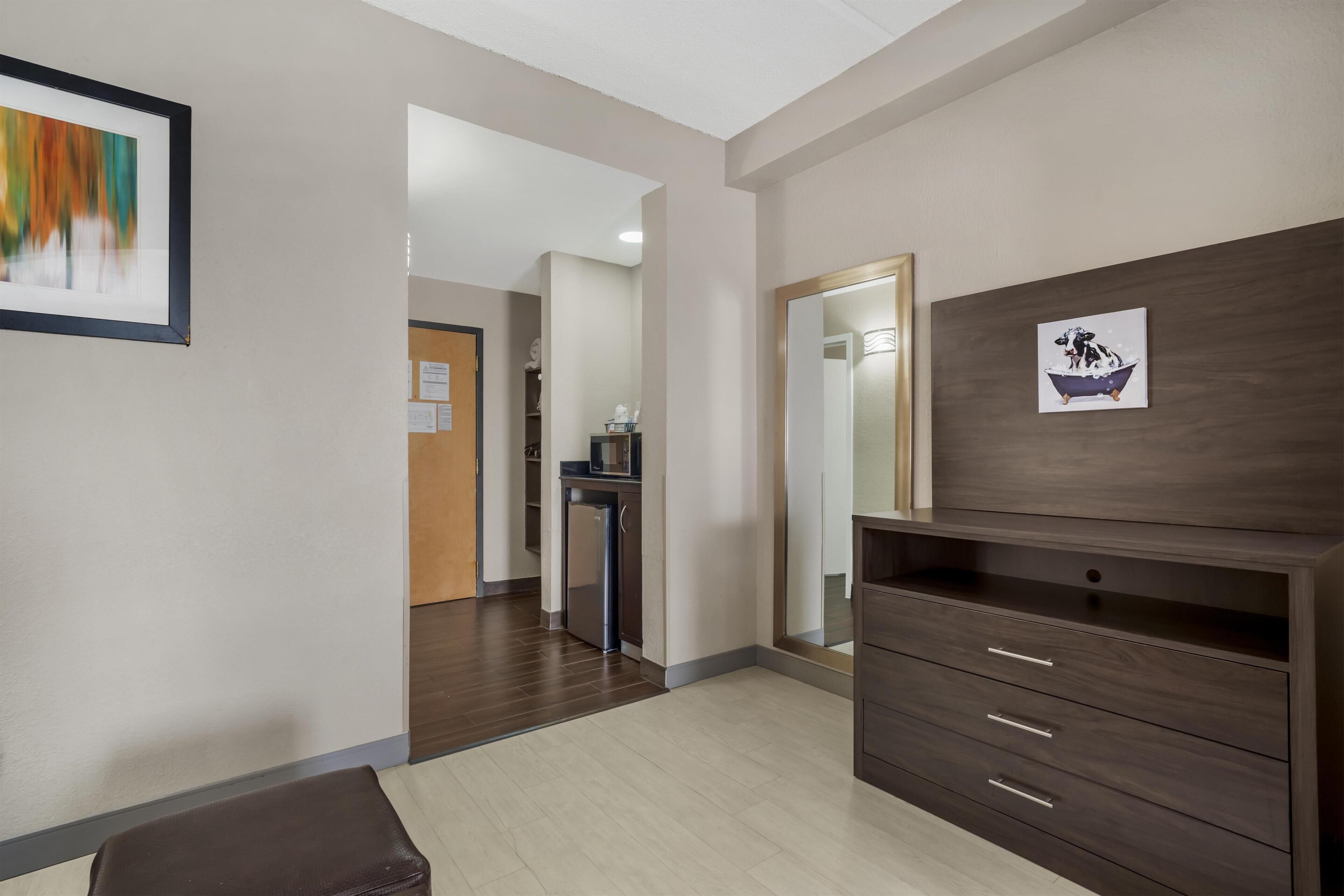 A hotel room with a wooden dresser, a mirror, a framed picture, and a door leading to a hallway.