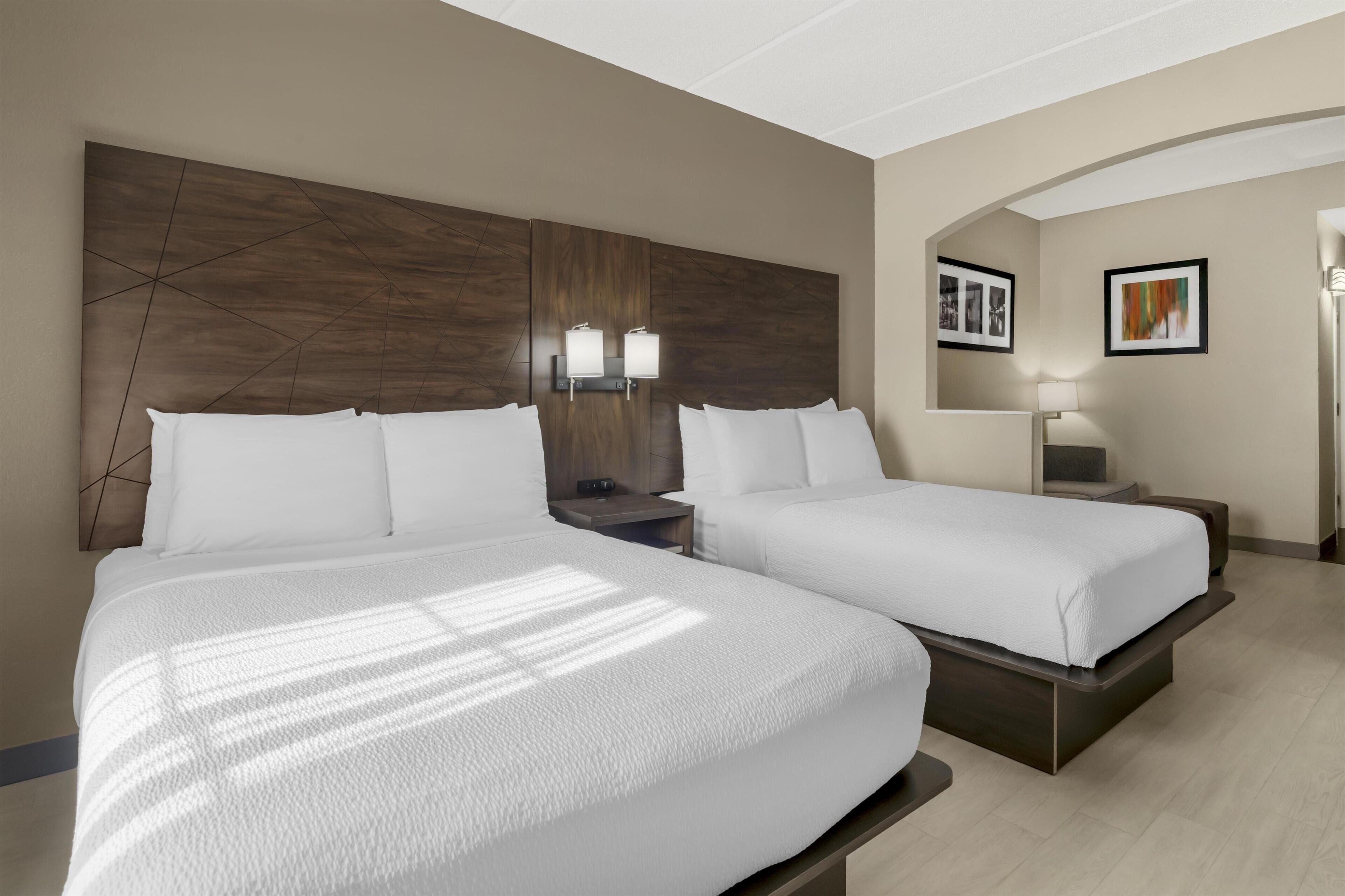 A hotel room with two beds, a wooden headboard, bedside lamps, a nightstand, and framed pictures on the wall.