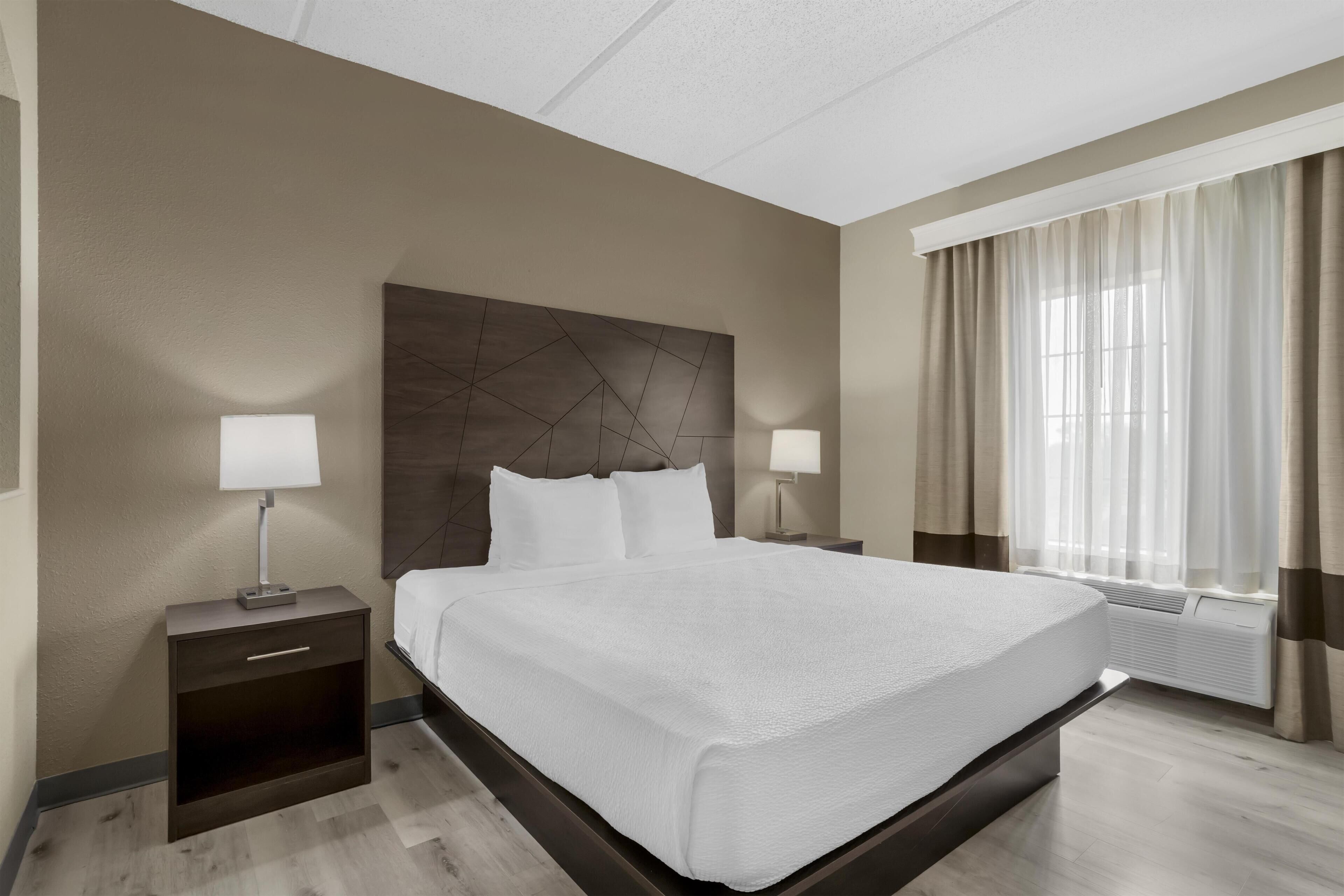 A hotel room with a large bed, two bedside tables with lamps, a modern headboard, and a large window with curtains.