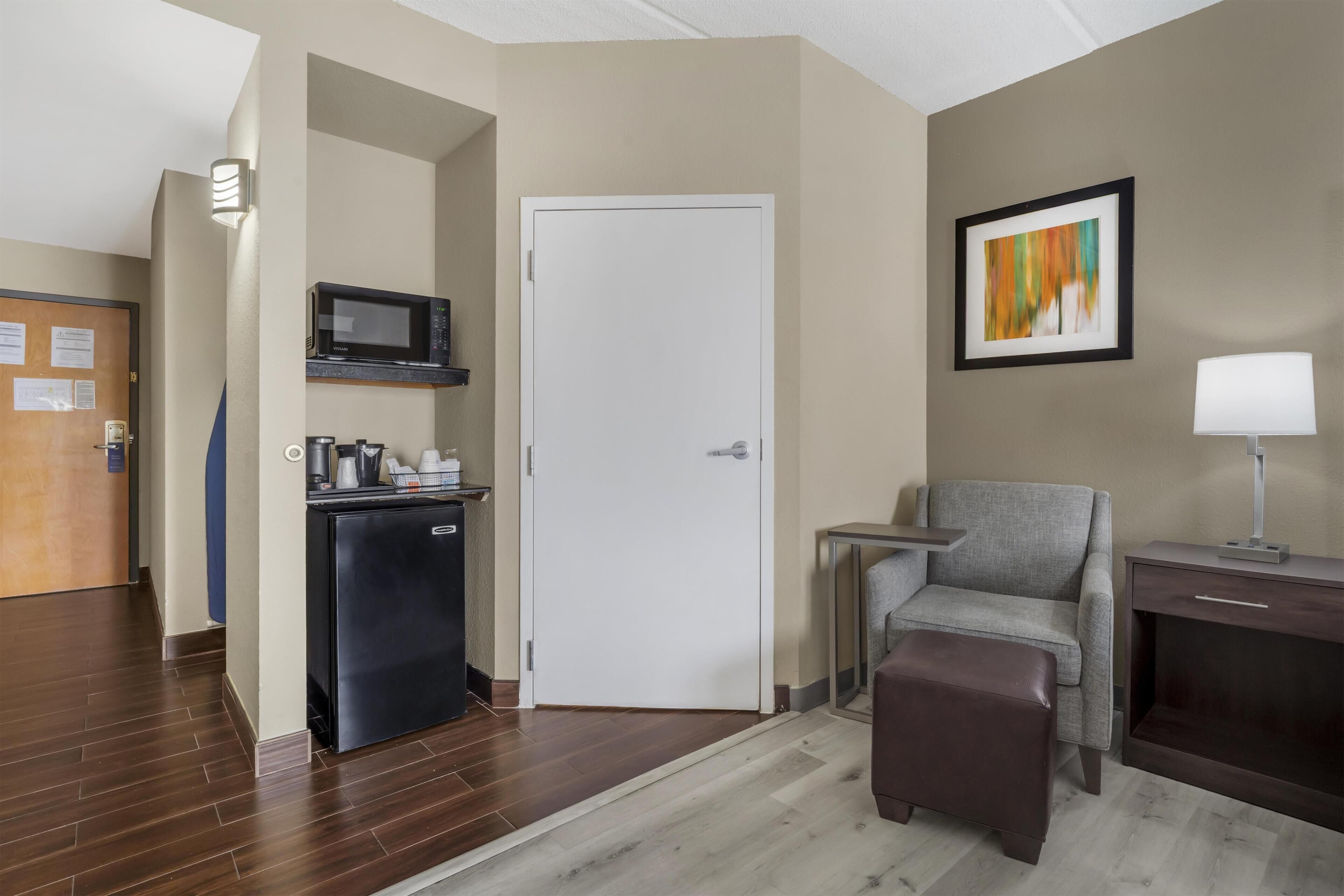Habitación de hotel con kitchenette, una puerta, un sillón gris, una mesita, una lámpara y un cuadro en la pared.