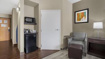Habitación de hotel con kitchenette, una puerta, un sillón gris, una mesita, una lámpara y un cuadro en la pared.
