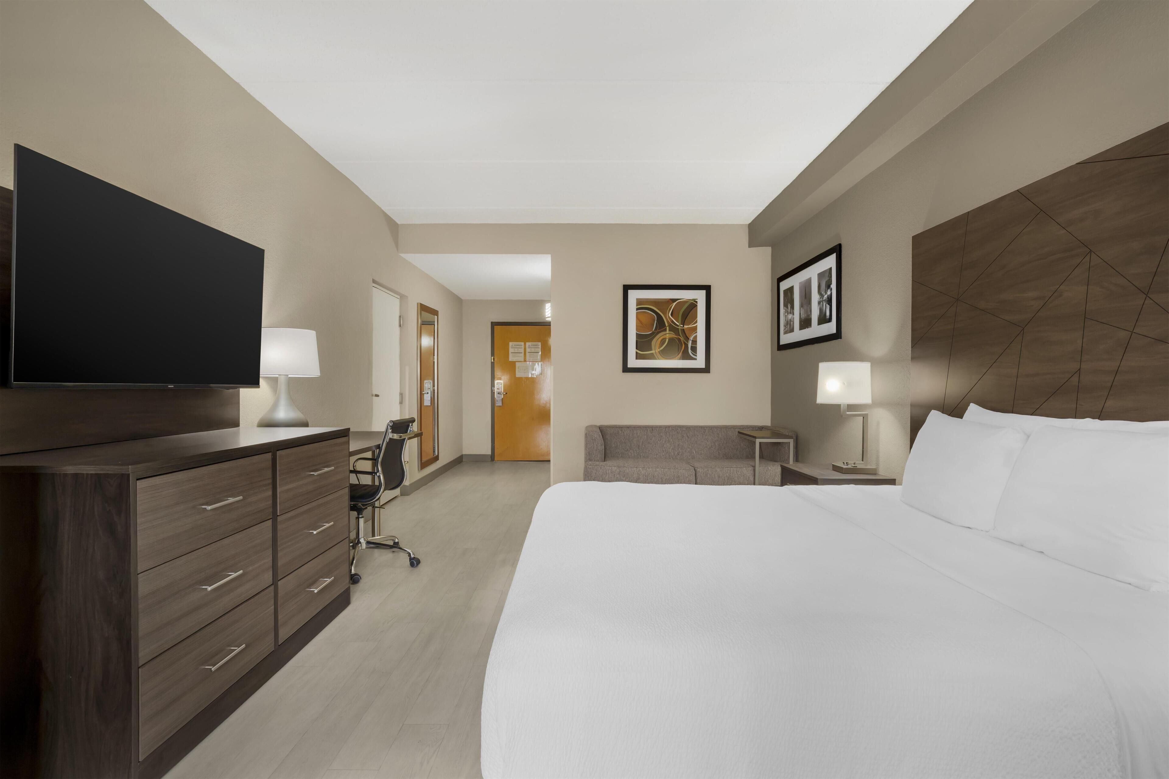Habitación de hotel con una cama grande, un televisor de pantalla plana, un escritorio con lámpara, una silla y una puerta que da a otra habitación.