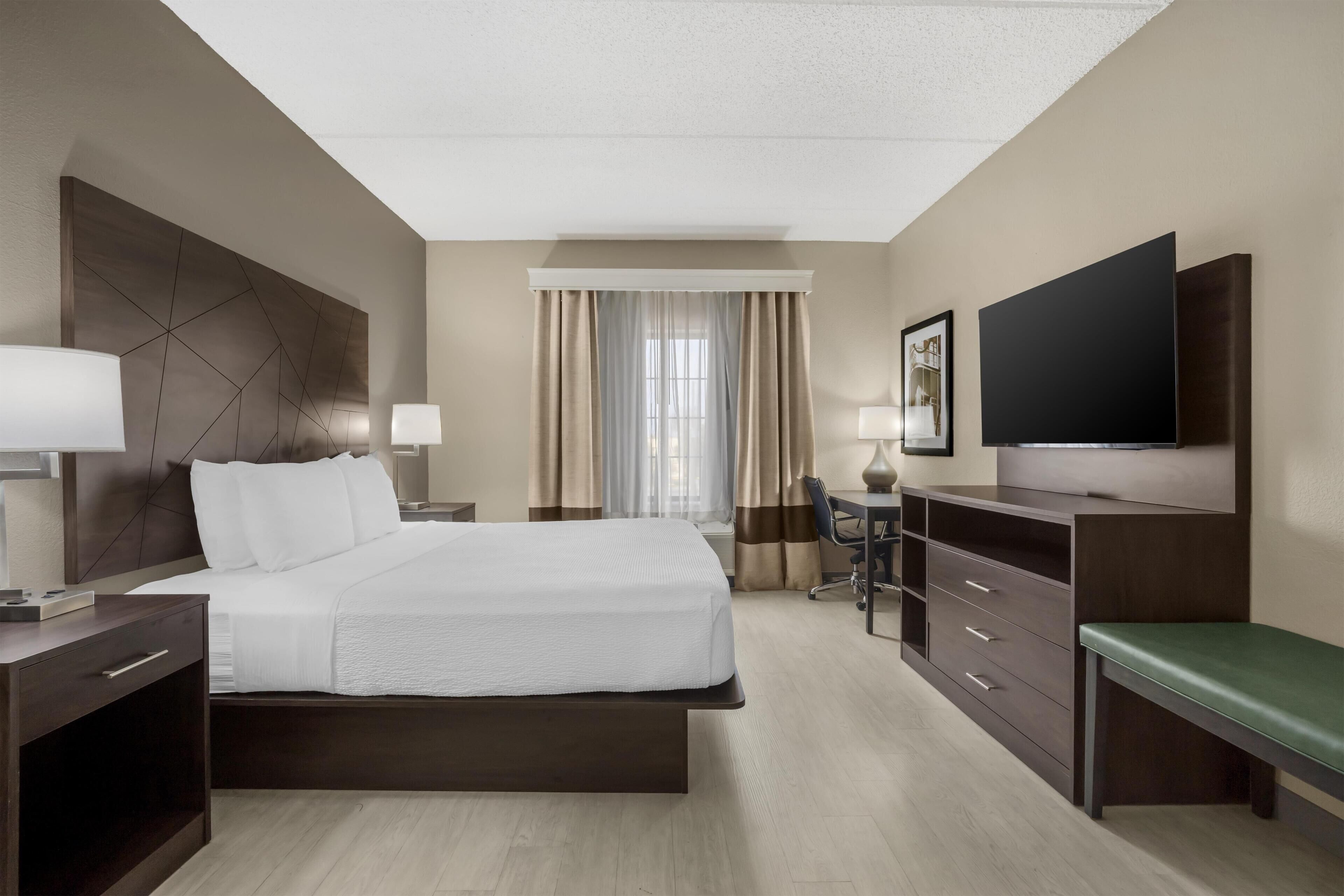 Habitación de hotel con cama, escritorio, silla, televisor y ventana con cortinas.