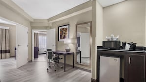 A hotel room with a desk, chair, kitchenette, and a door leading to a bathroom.