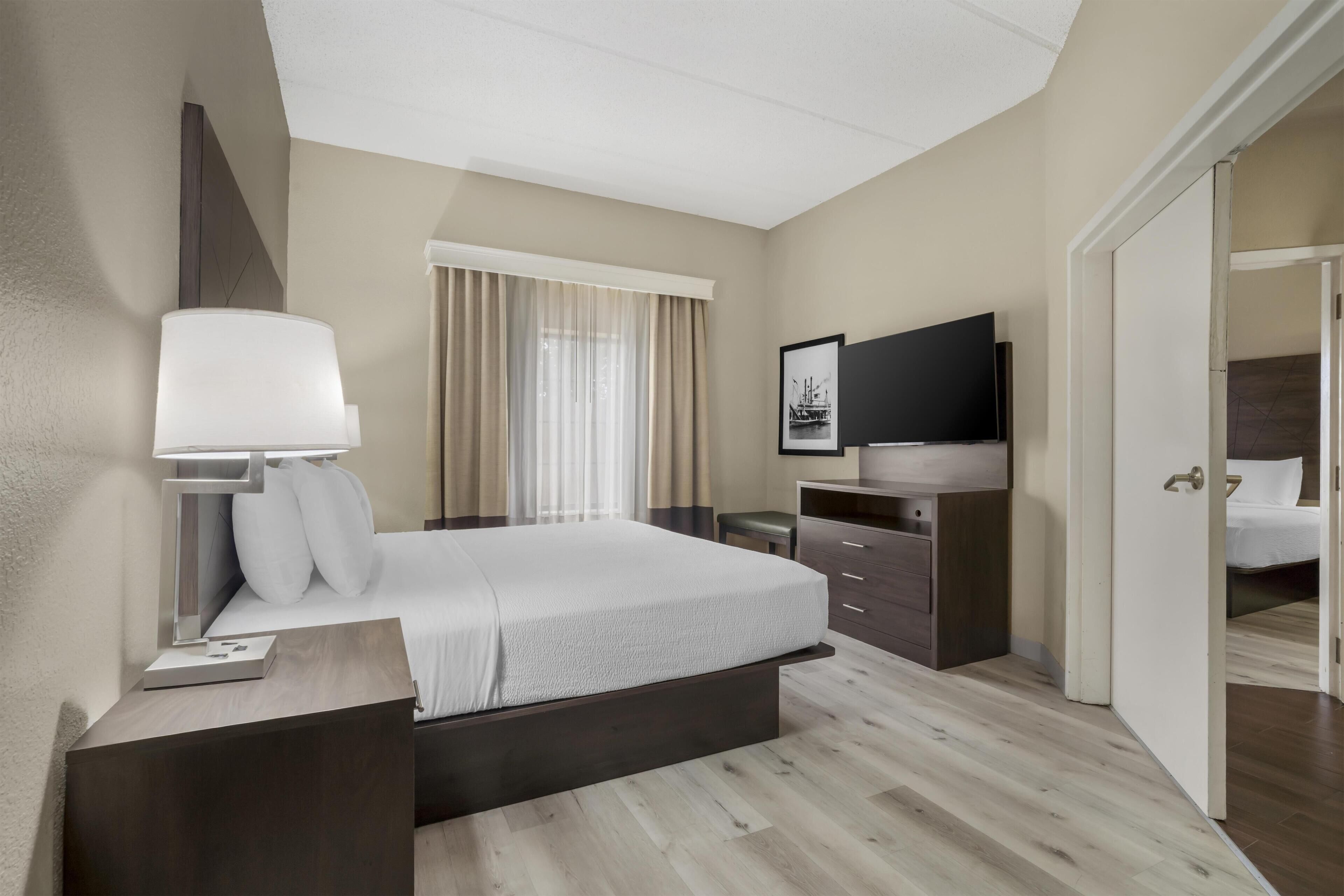 Habitación de hotel con cama, mesita de noche, lámpara, televisión y puerta que da a otra estancia.