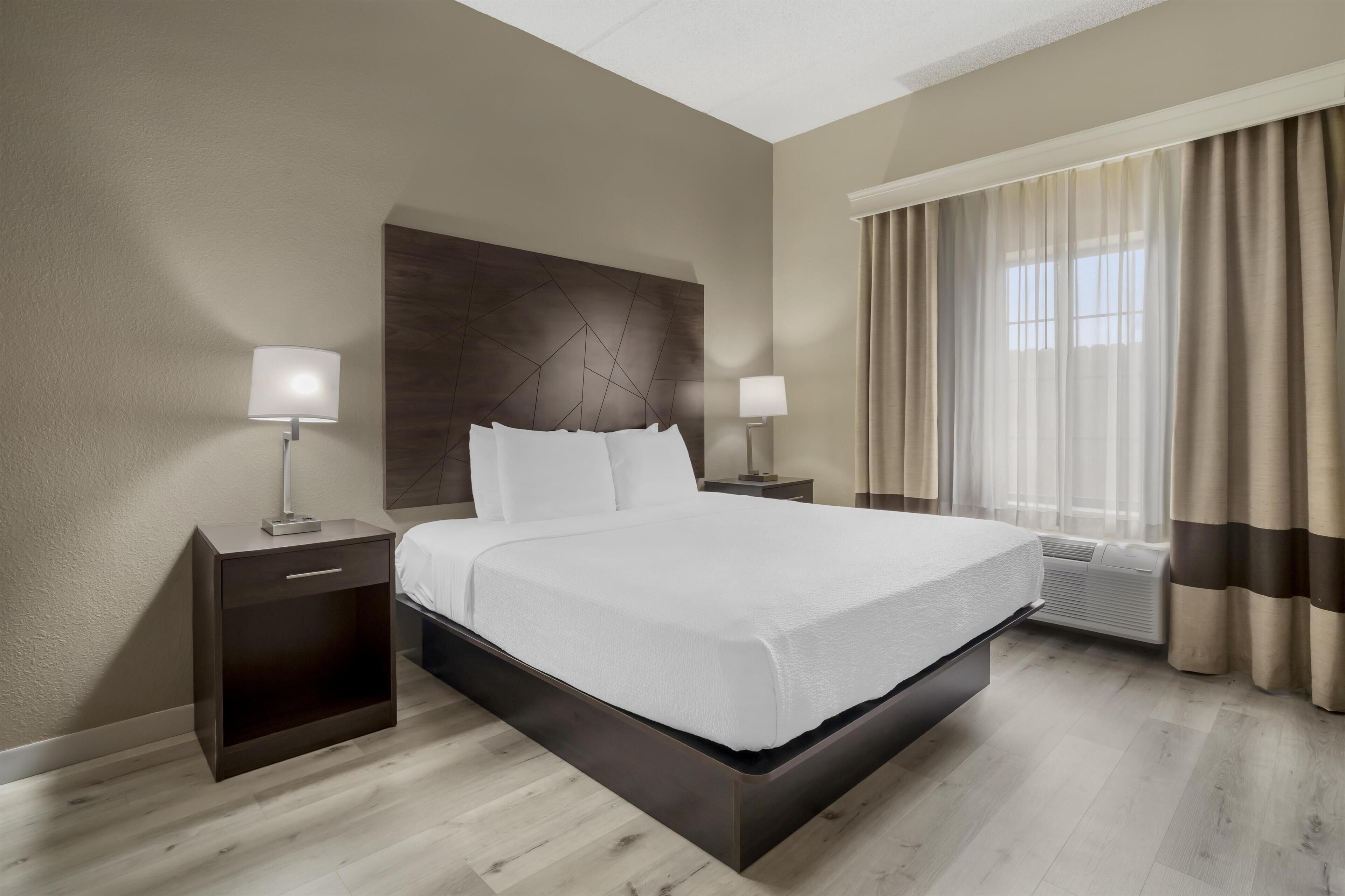 Hotelkamer met een groot bed, twee nachtkastjes met lampen, een hoofdbord, een raam met gordijnen en een airconditioning.