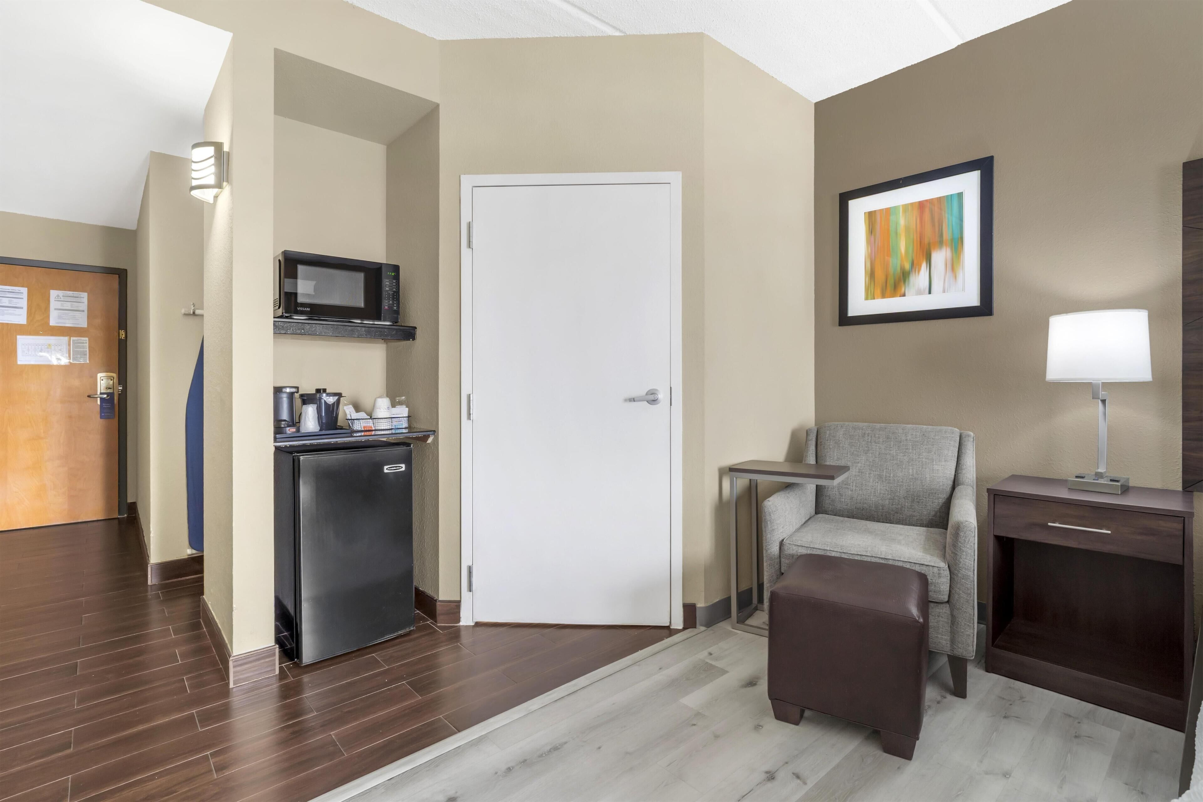 A hotel room with a kitchenette, a door, a chair, a small table, a lamp, and a painting on the wall.