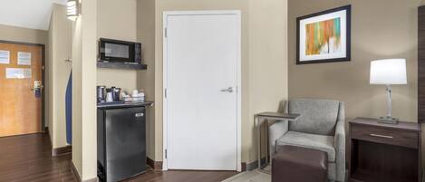 A hotel room with a kitchenette, a door, a chair, a small table, a lamp, and a painting on the wall.