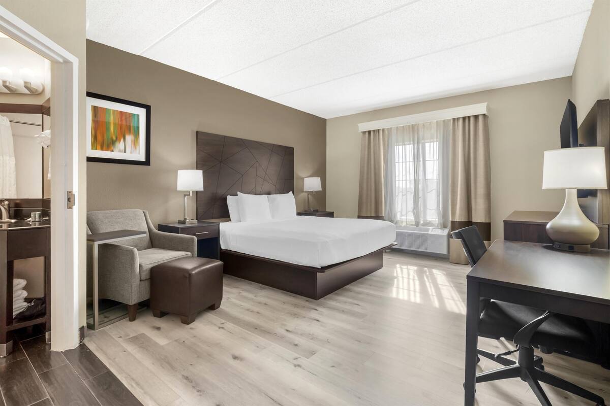 Une chambre d’hôtel comprenant un lit, un bureau, une chaise, un canapé, une table de chevet et une fenêtre avec des rideaux.