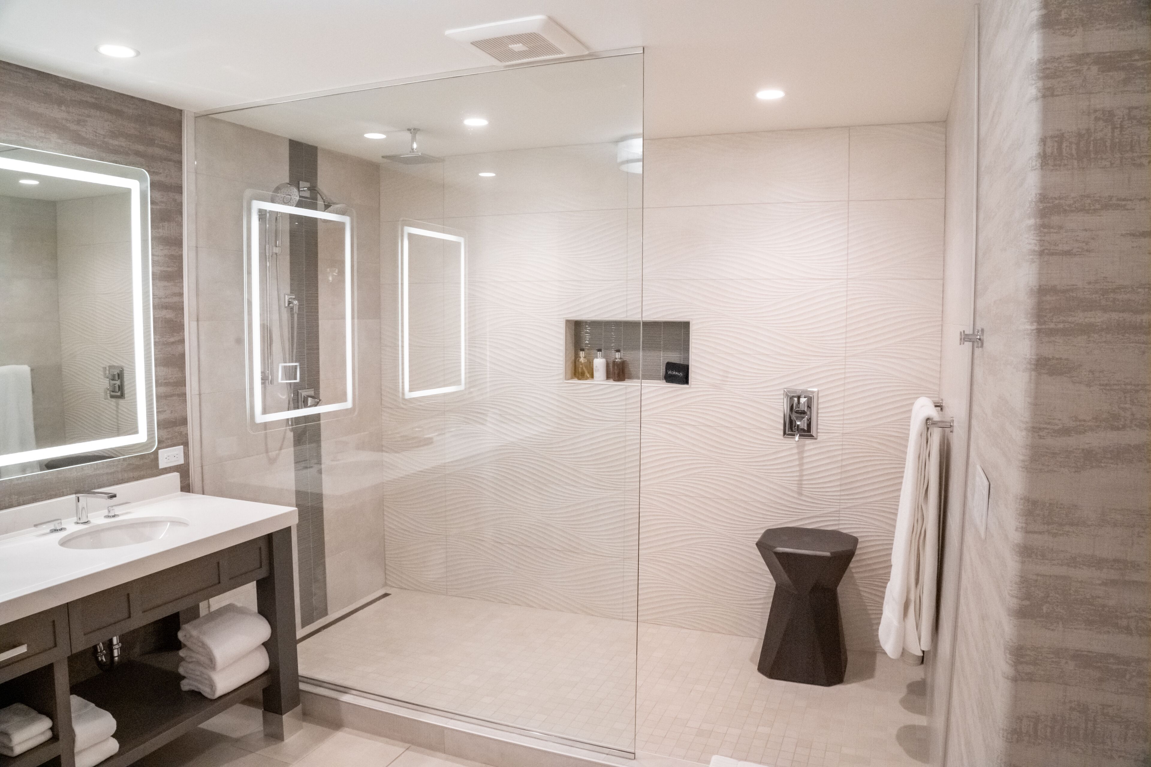 Un baño moderno con ducha envuelta en vidrio, un espejo grande y una tocador con lavabo y toallero.
