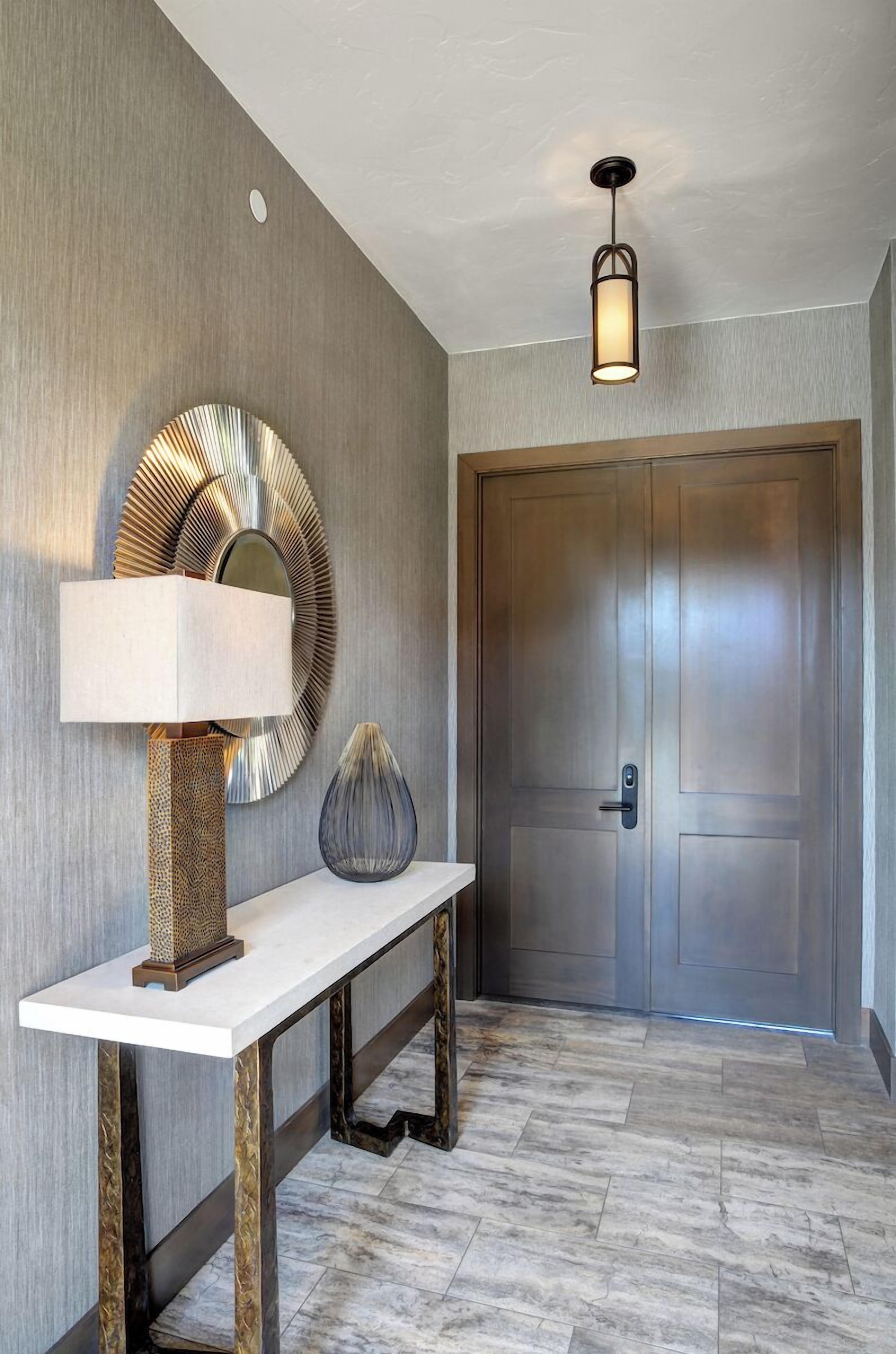 A hallway with a wooden door, a white console table with a lamp, a decorative mirror, and a hanging light fixture.