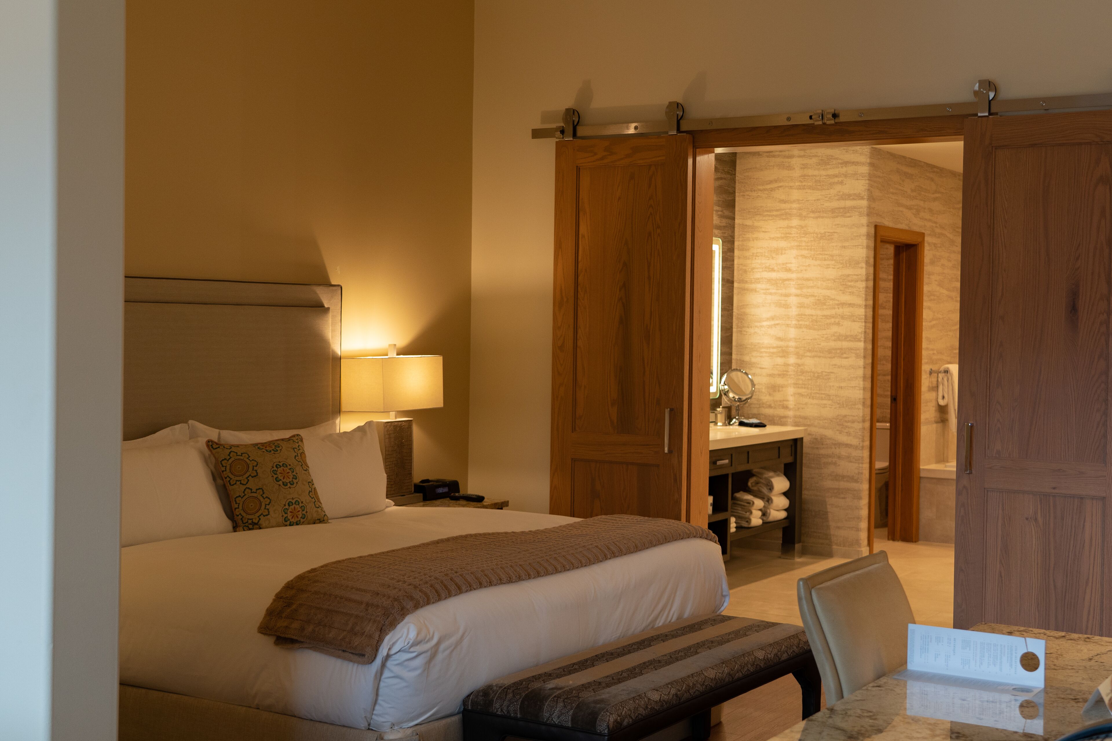 A hotel room with a large bed, bedside lamp, wooden wardrobe, and a bathroom visible through an open door.