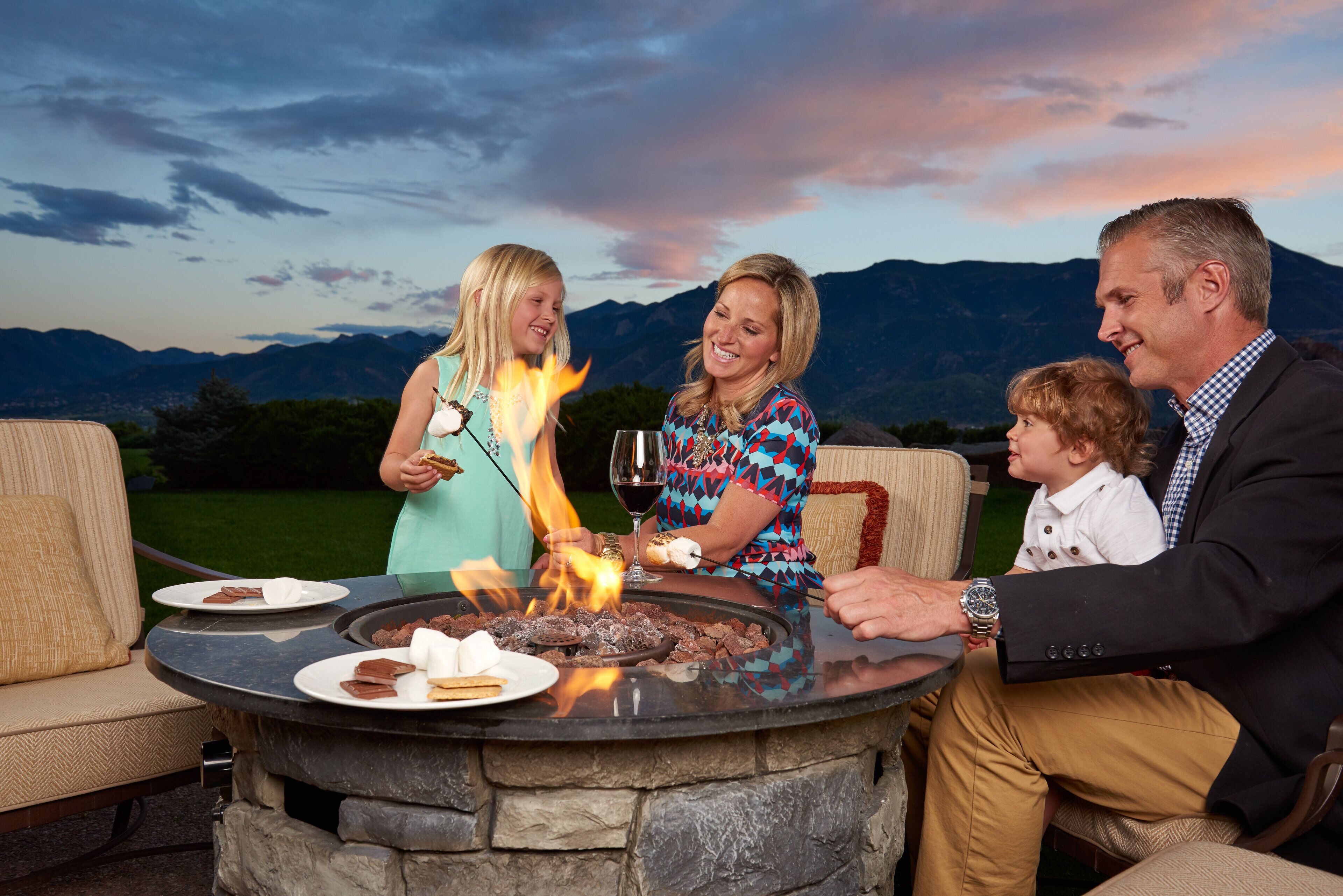 A family gathered around a stone fire pit with food and drinks, set against a mountainous backdrop.