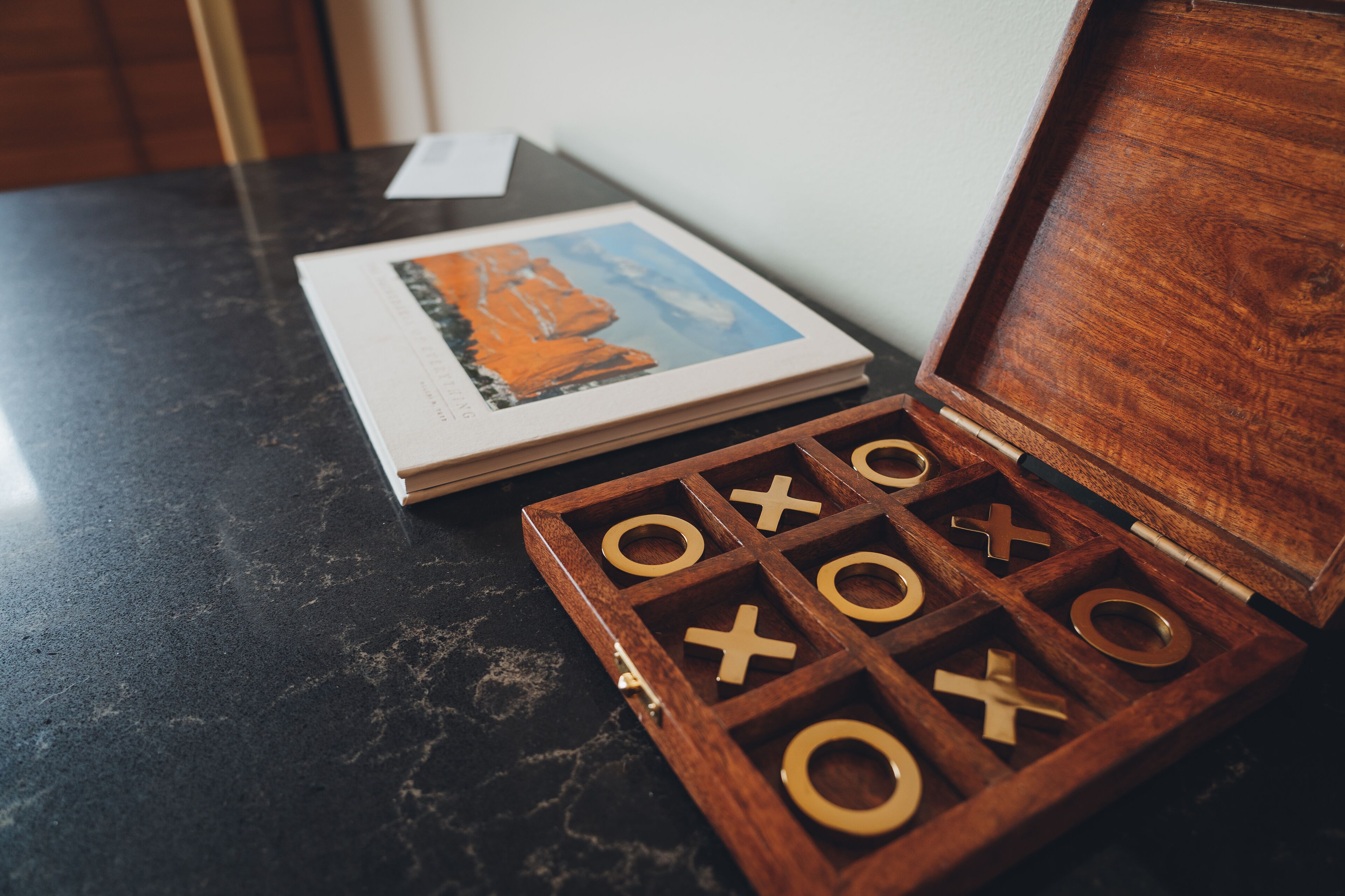 A wooden tic-tac-toe game set on a dark marble table with a book and a framed picture in the background.