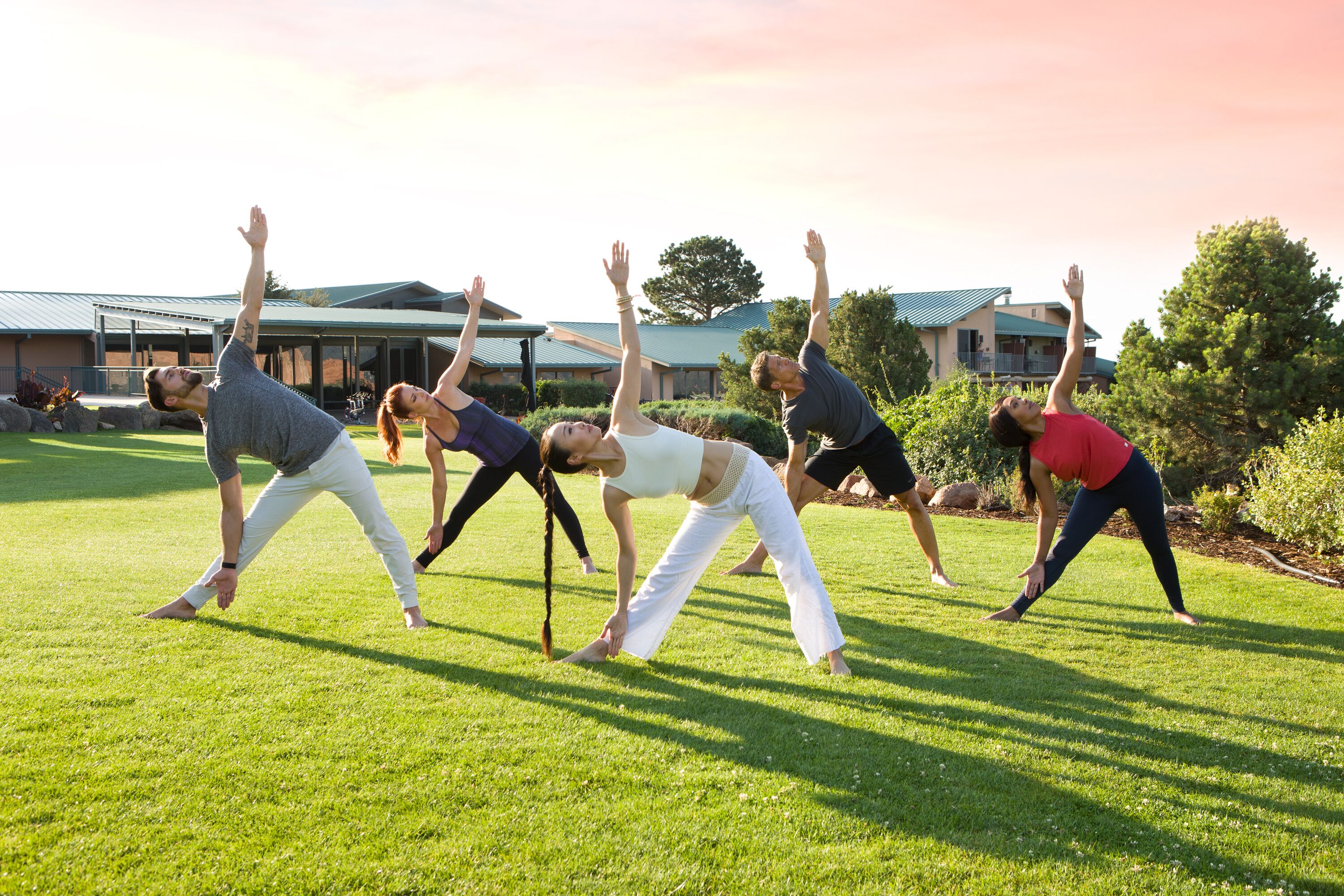 A group of people performing a yoga stretch outdoors on a grassy field with buildings in the background.