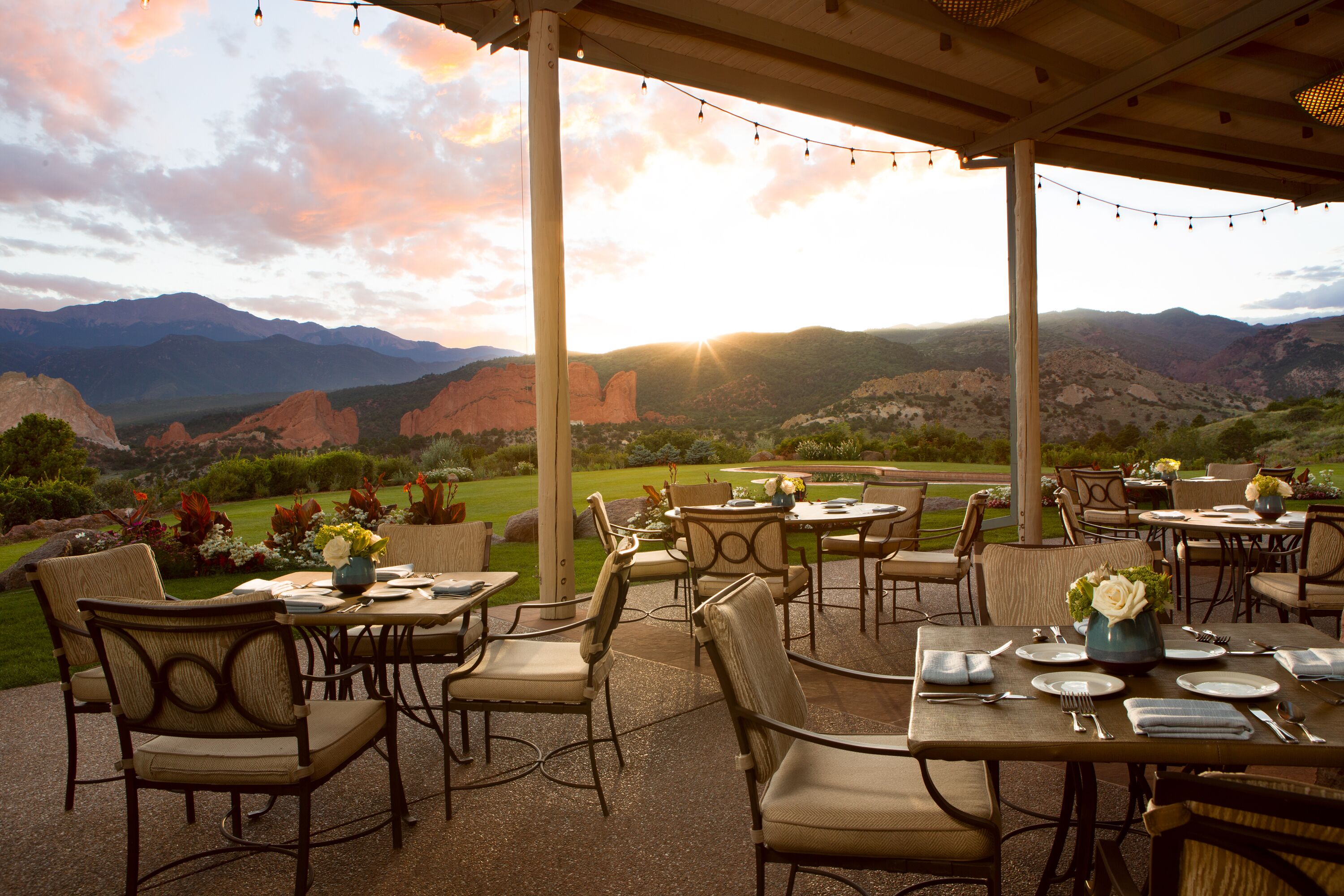Un patio al aire libre con mesas y sillas, con montañas y un atardecer de fondo.