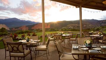 Un patio al aire libre con mesas y sillas, con montañas y un atardecer de fondo.