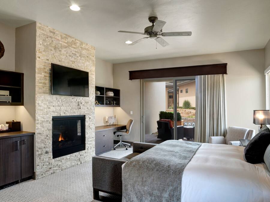 Une chambre d’hôtel moderne avec un mur d’accent en pierre, une cheminée, un téléviseur à écran plat, un bureau avec une chaise, un lit avec un grand oreiller et un ventilateur de plafond.