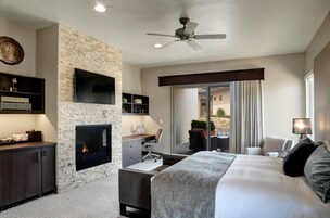 A modern hotel room with a stone accent wall, a fireplace, a flat-screen TV, a desk with a chair, a bed with a large pillow, and a ceiling fan.