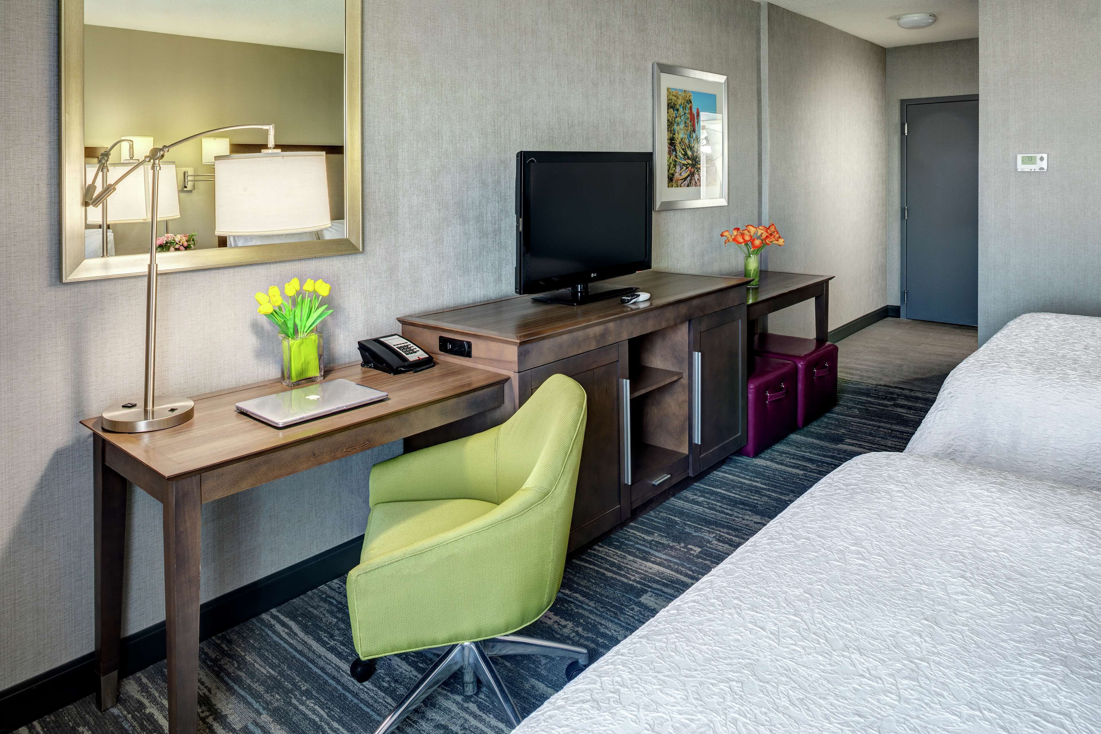 A hotel room with a bed, a desk with a computer, a chair, a TV, a lamp, a vase with flowers, and a door.