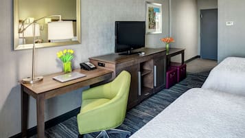 A hotel room with a bed, a desk with a computer, a chair, a TV, a lamp, a vase with flowers, and a door.