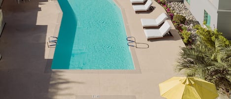 Piscina con tumbonas y sombrillas, rodeada de vegetación y un edificio.
