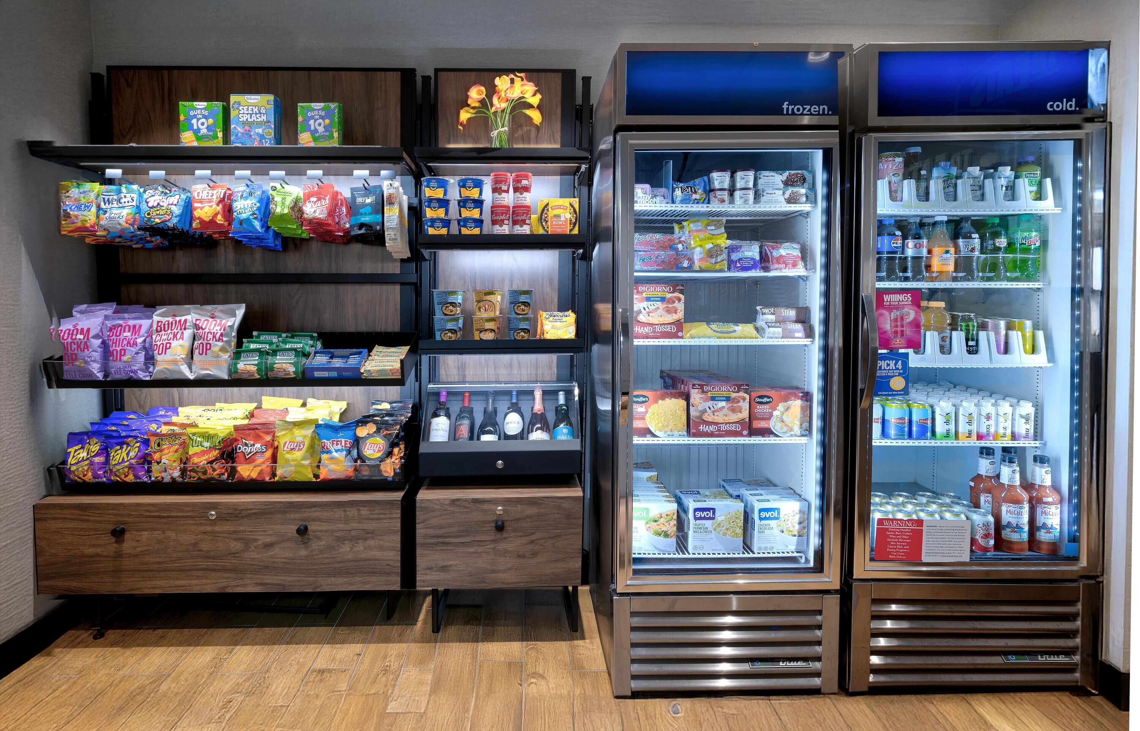 A vending machine stocked with snacks and beverages, a refrigerated section with frozen and cold items, and a wooden shelf with various packaged goods.