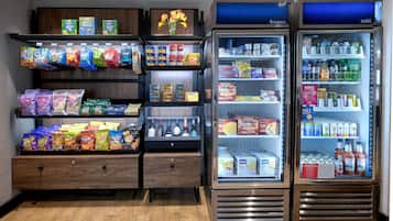 A vending machine stocked with snacks and beverages, a refrigerated section with frozen and cold items, and a wooden shelf with various packaged goods.
