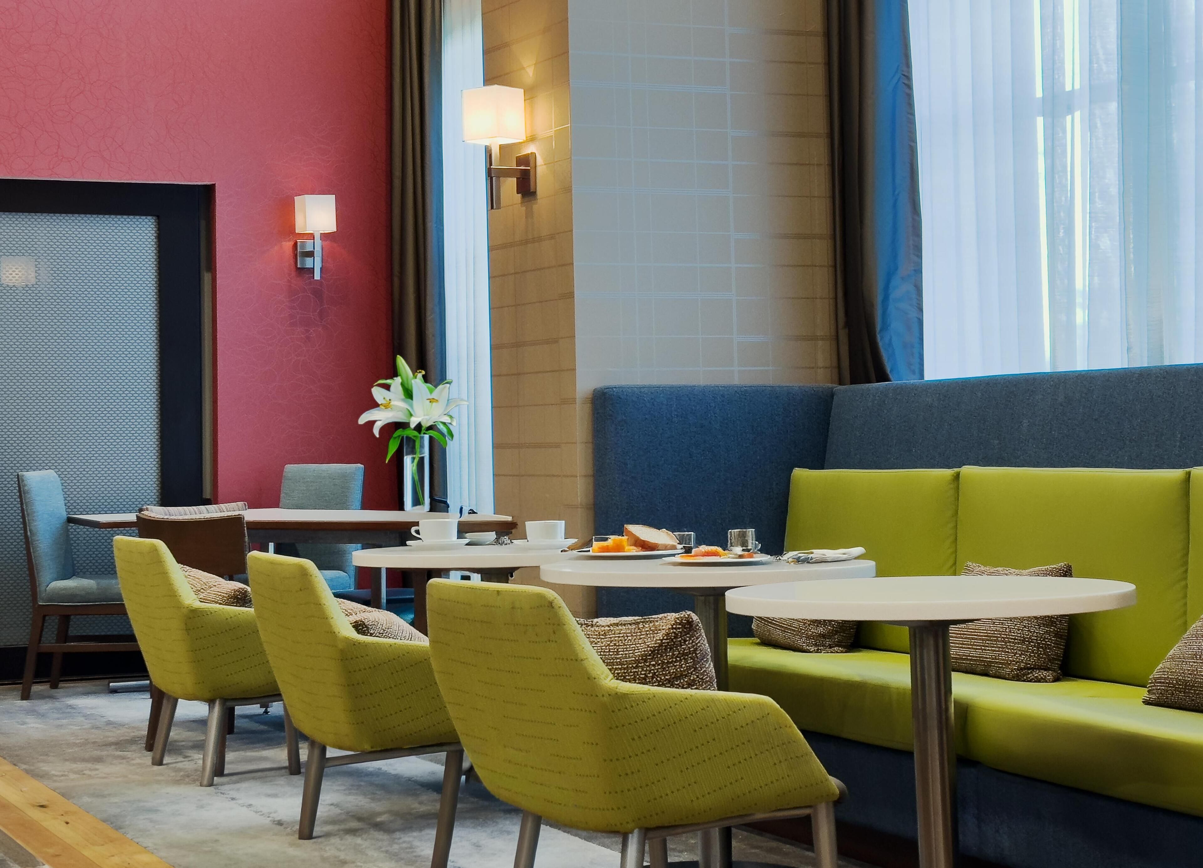 A modern lounge area with green and blue seating, a small table with a vase of flowers, and a red accent wall.