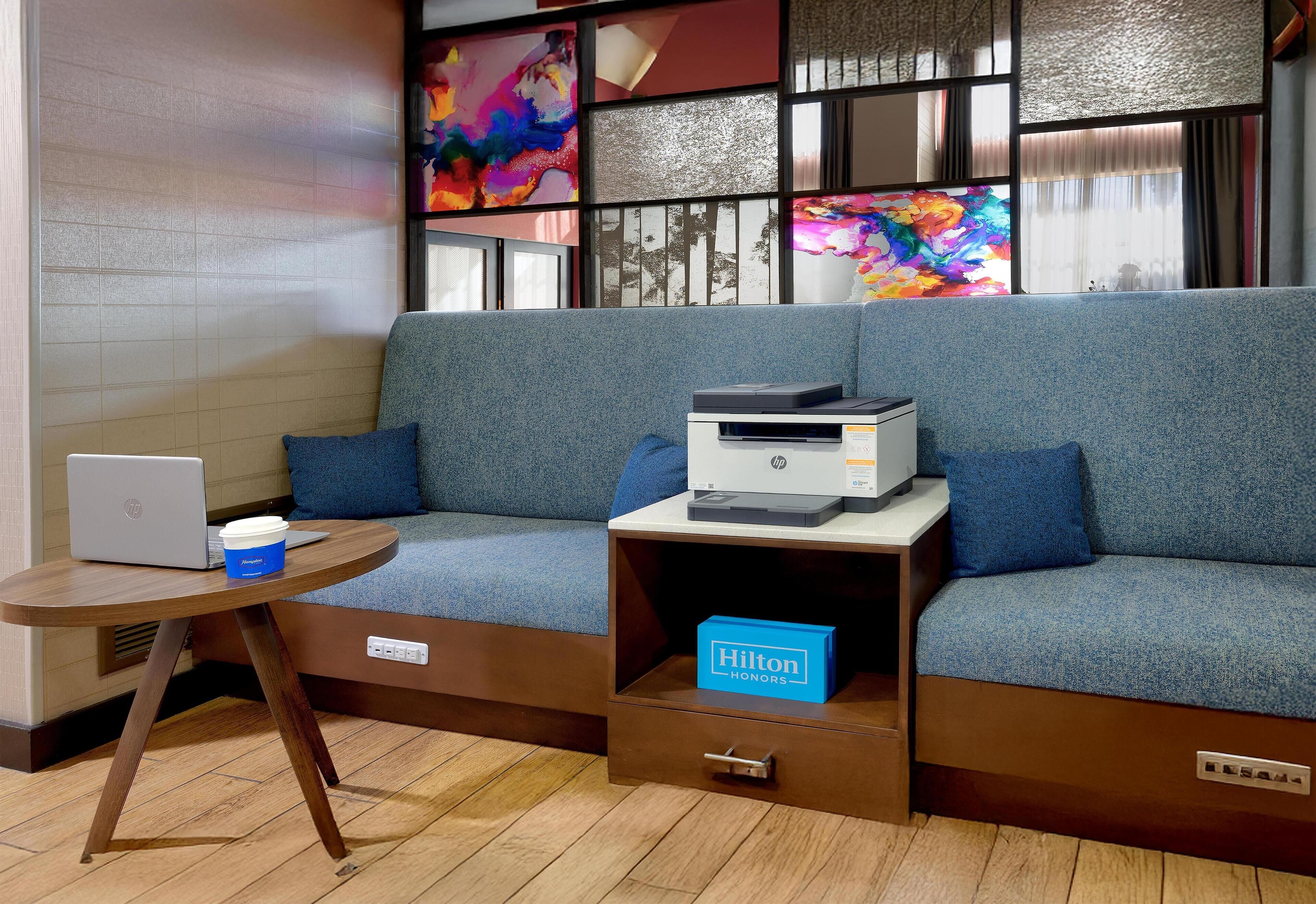 Une chambre d’hôtel moderne avec un canapé gris, une petite table en bois sur laquelle se trouvent un ordinateur portable et une tasse de café, et une imprimante posée sur une table d’appoint.