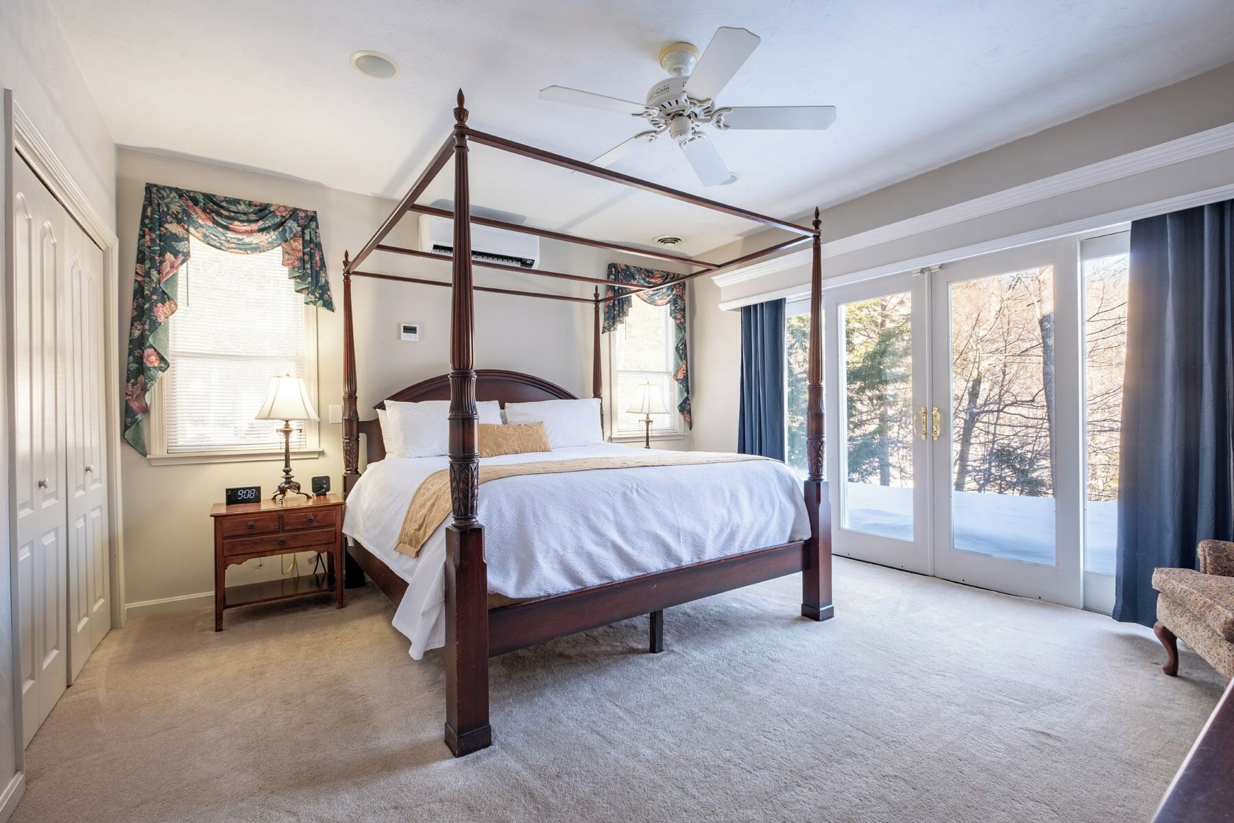 A four-poster bed with a ceiling fan, a nightstand with a lamp, and a view of trees through large windows.