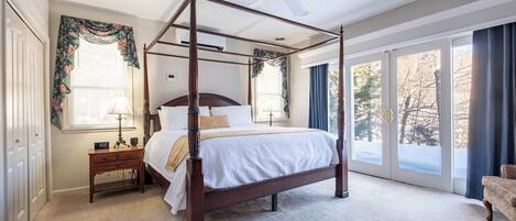 A four-poster bed with a ceiling fan, a nightstand with a lamp, and a view of trees through large windows.