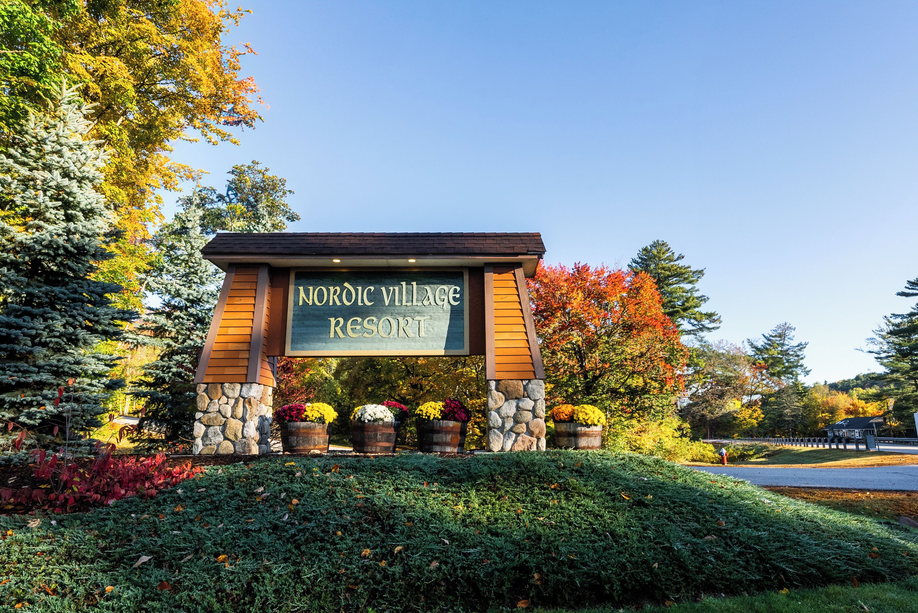 A sign for Nordic Village Resort with a stone and wood structure, surrounded by autumn foliage and potted plants.