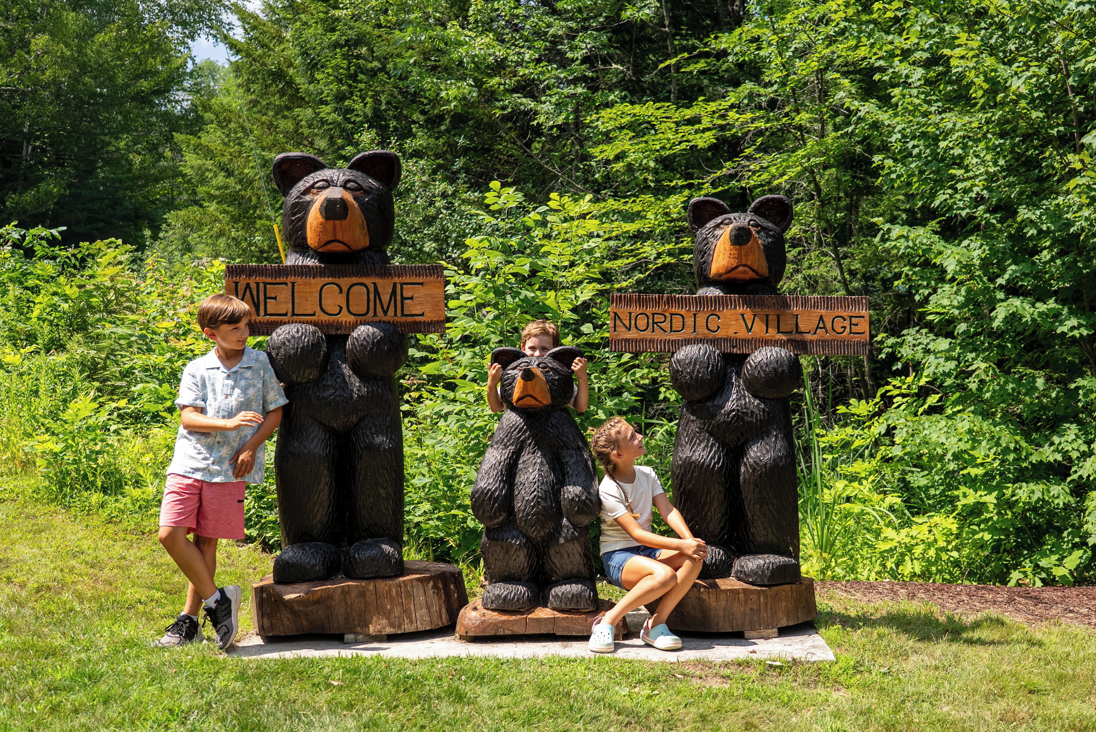 Three wooden bear sculptures with signs, one saying 'WELCOME' and the other 'NORDIC VILLAGE', set against a backdrop of greenery.