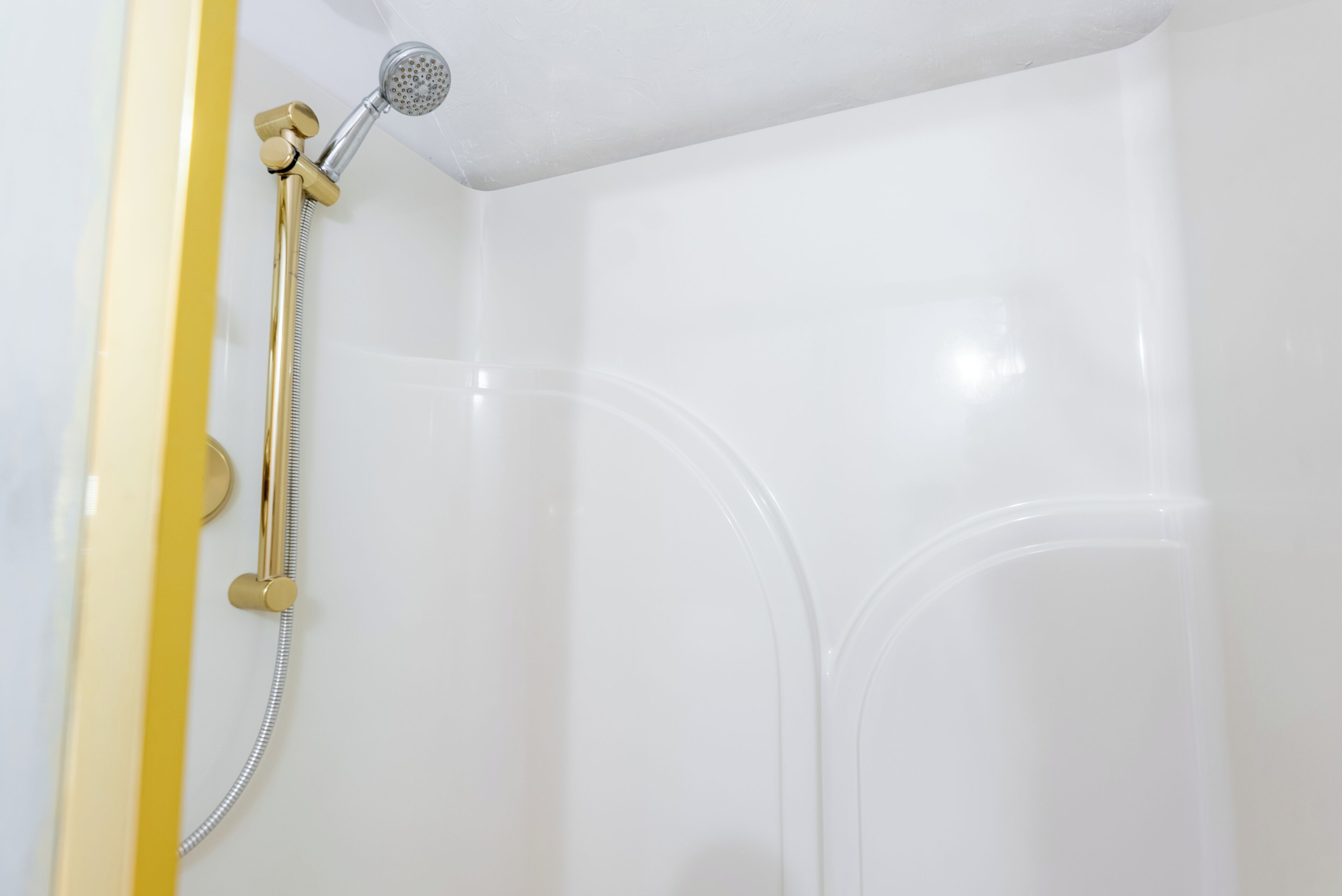 A shower with a gold showerhead and hose, white walls, and a yellow door frame.