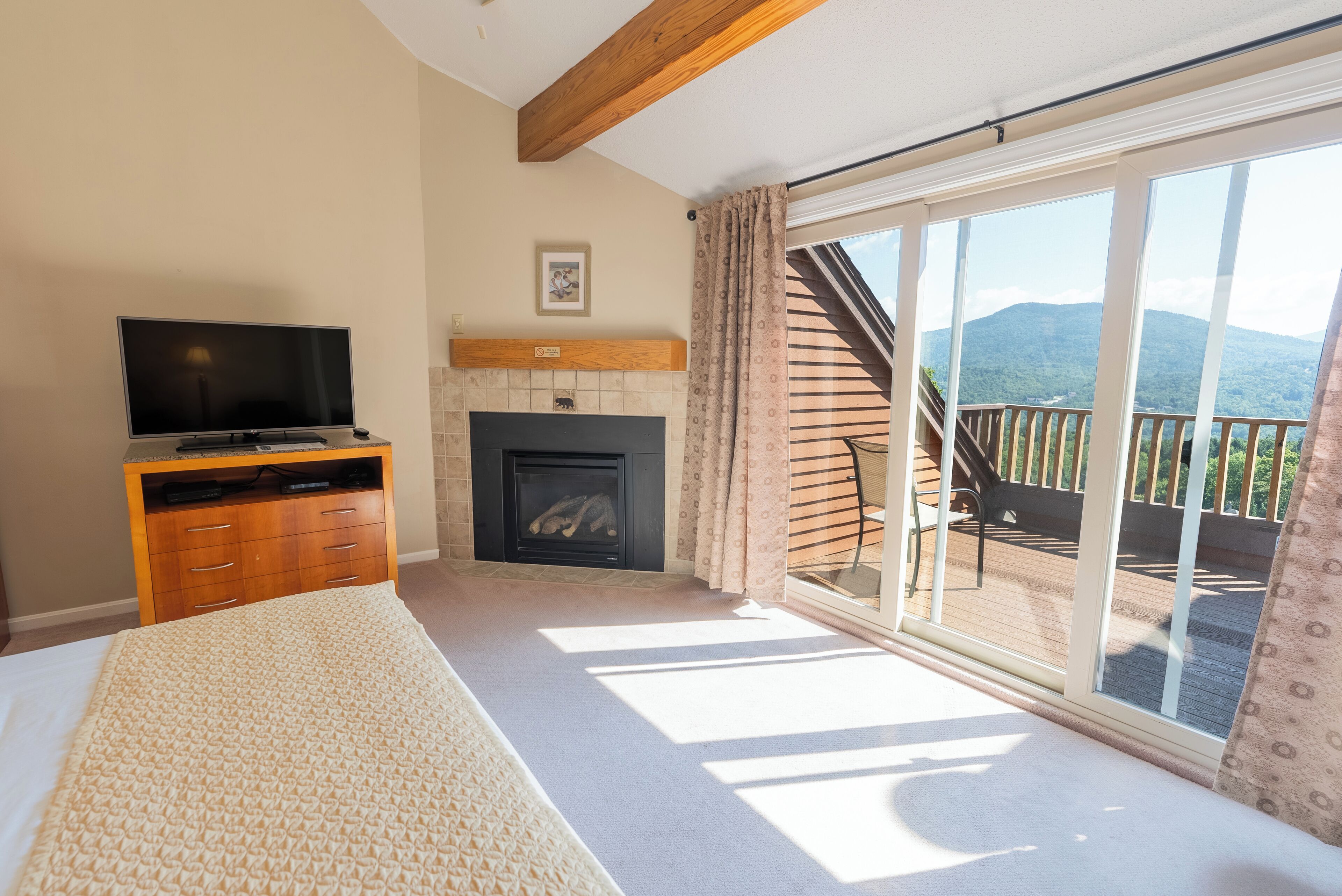 A bedroom with a large bed, a TV, a fireplace, and a balcony with a view of mountains.