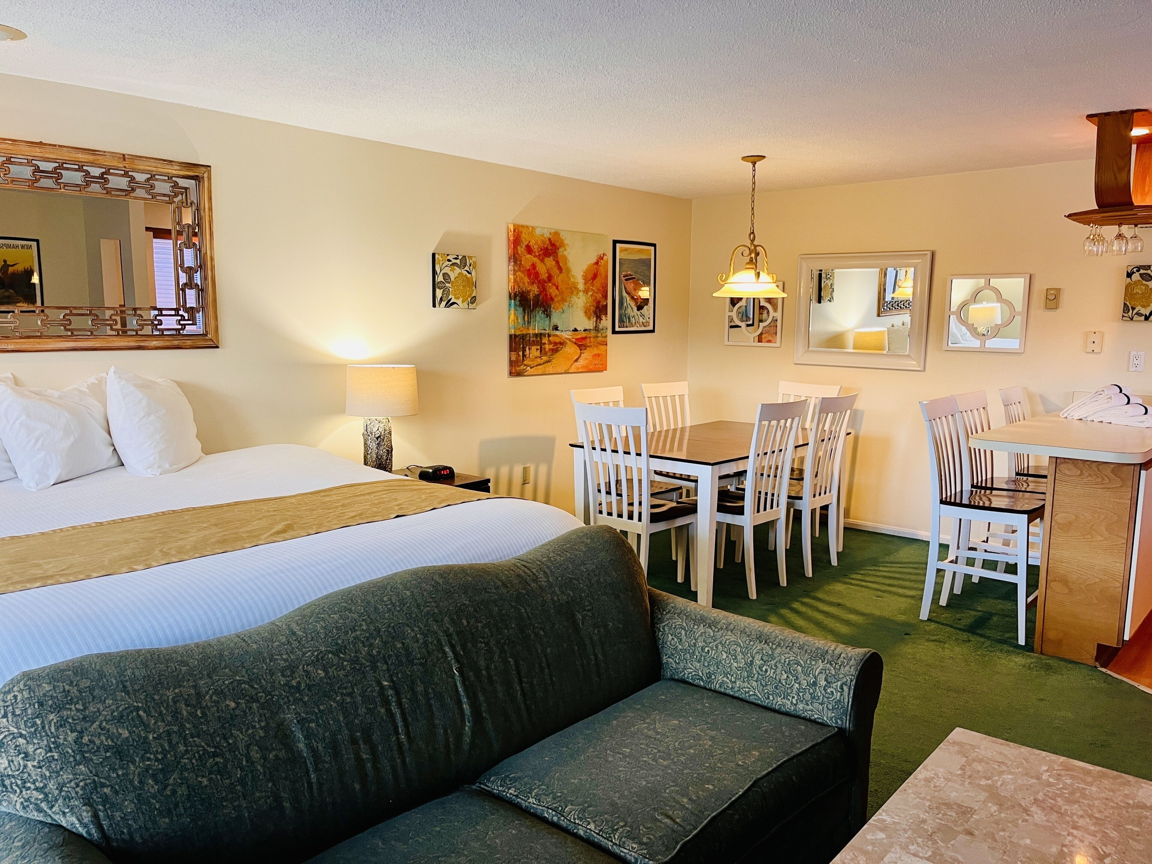 A hotel room with a large bed, a green sofa, a dining table with chairs, and a small kitchen area.