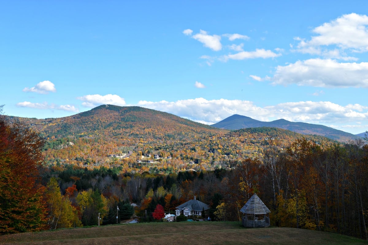 A rural landscape with a house and a gazebo in the foreground, surrounded by autumn-colored trees and hills in the background.