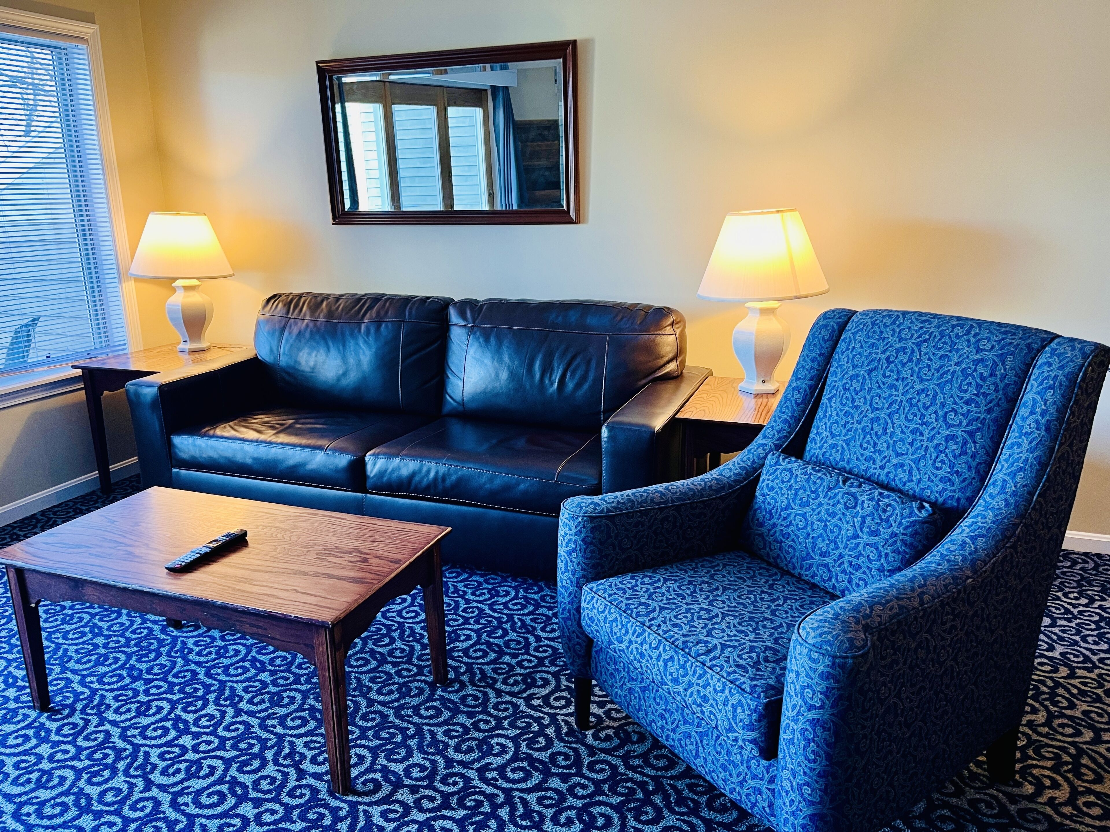 Ein Wohnzimmer mit einer schwarzen Ledersofa, einem blauen Ohrensessel, einem Holztisch und zwei Lampen auf Beistelltischen.