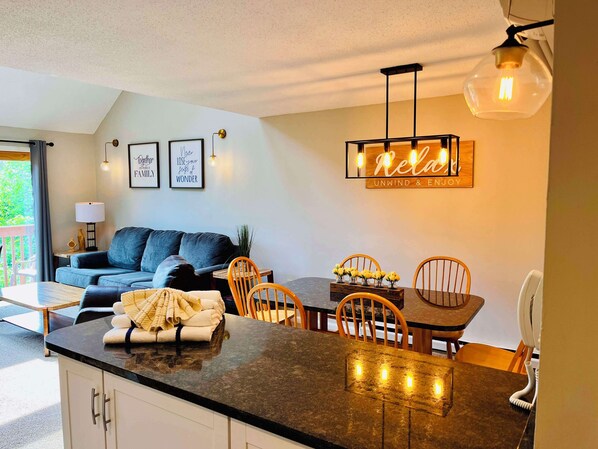 A modern kitchen with a dining area, featuring a kitchen island, wooden chairs, and a table with a neon sign.