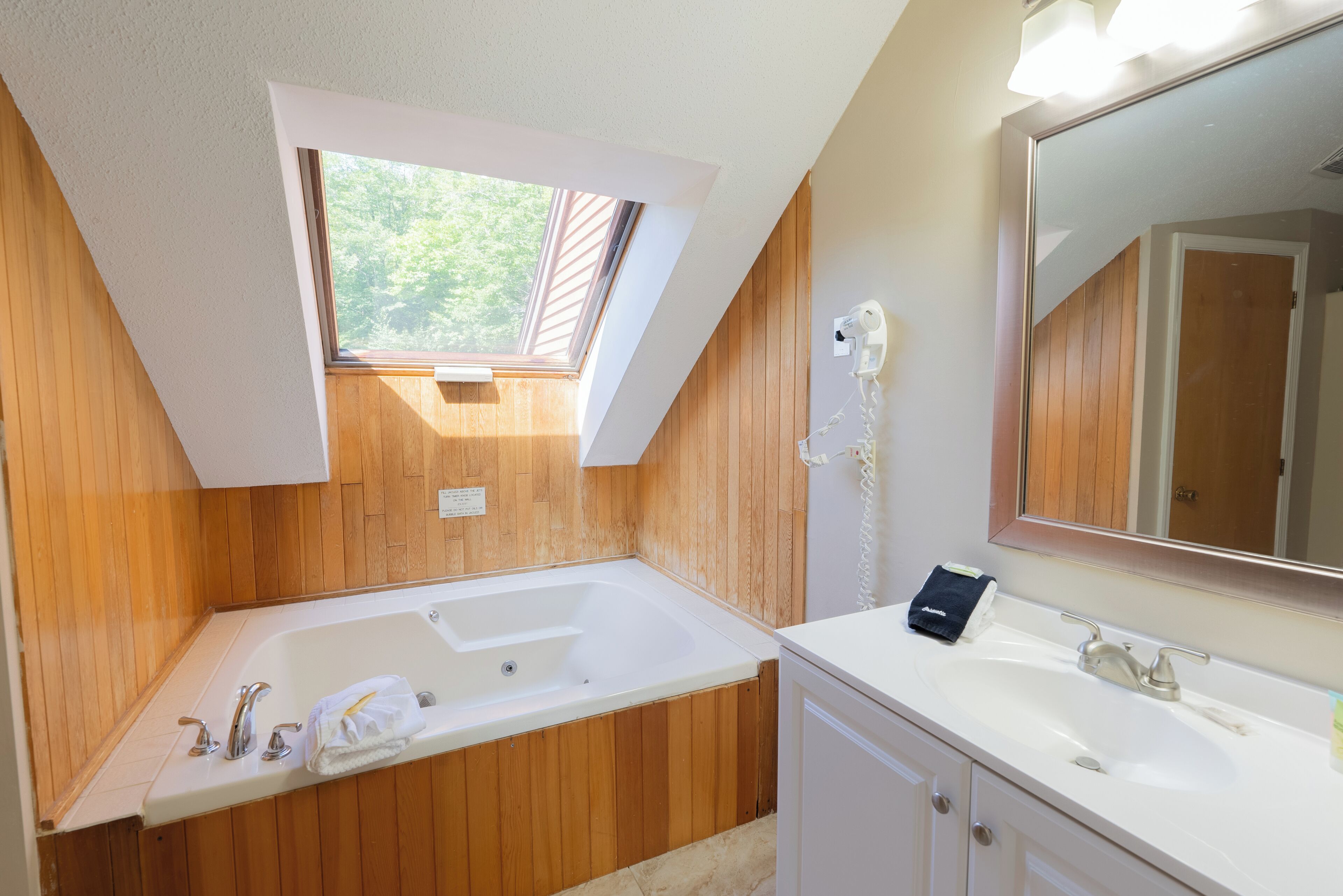 A bathroom with a wooden wall, a skylight, a white bathtub, a sink, and a mirror.