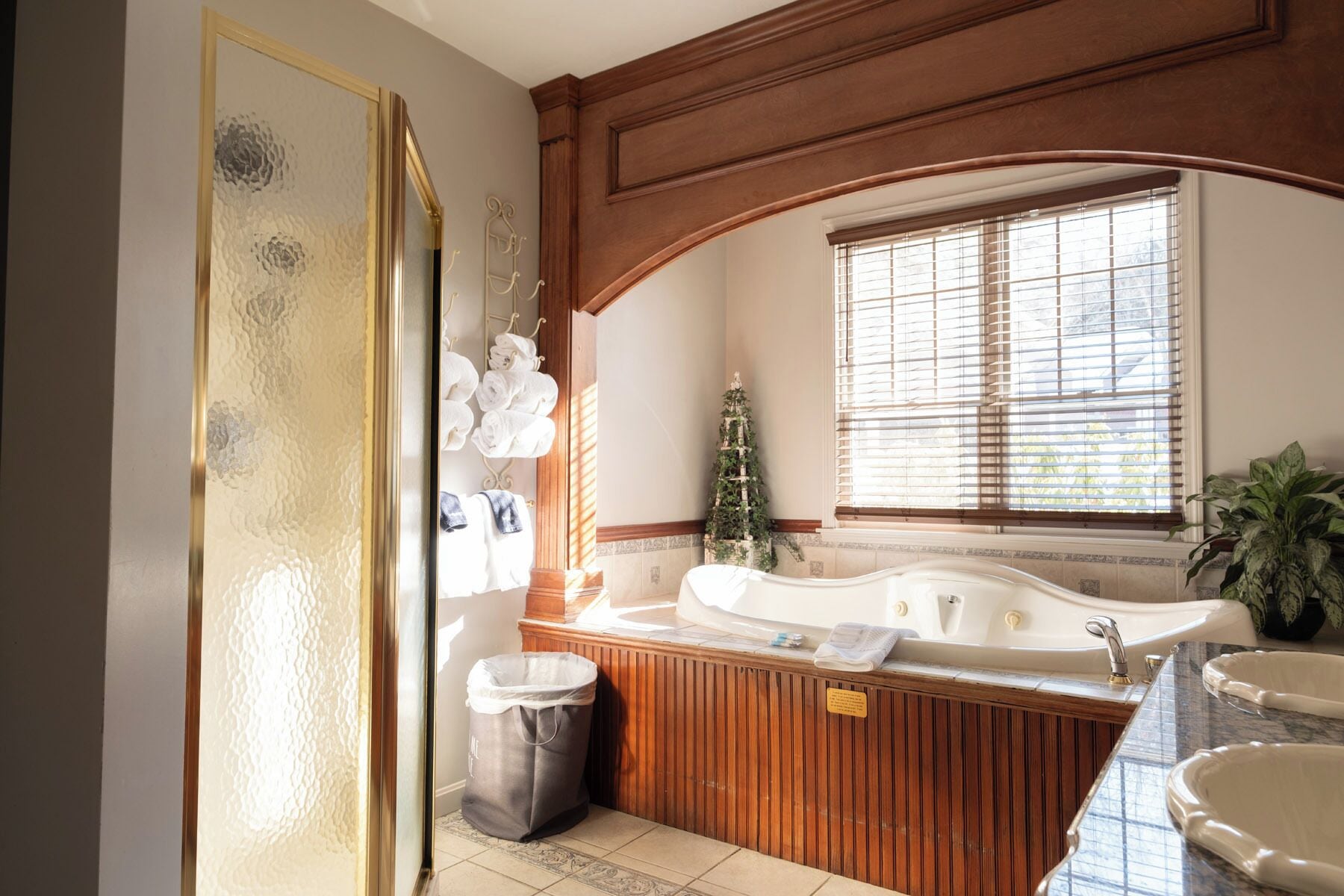A bathroom with a large bathtub, a wooden vanity, a window with blinds, and a towel rack with towels.